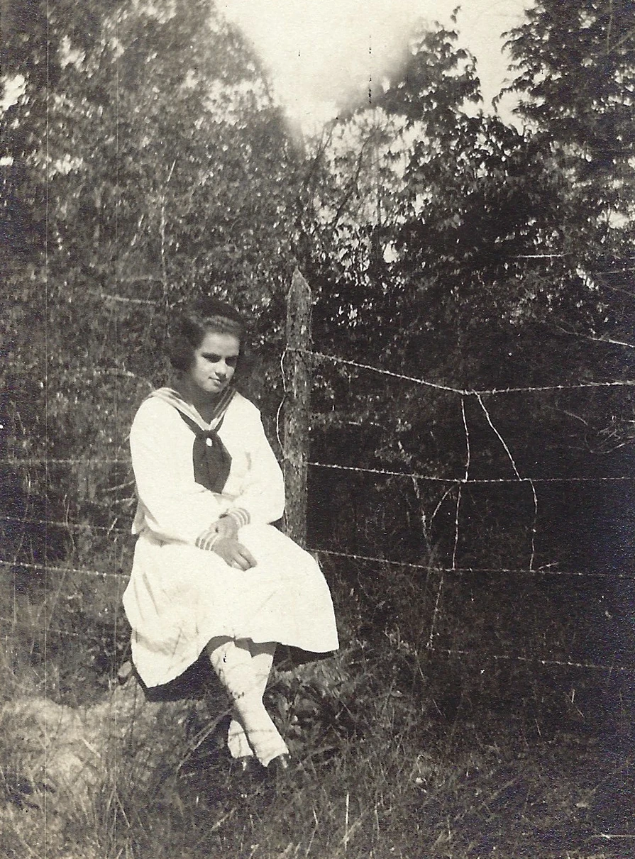 Mama Percy (Catherine Fleming) as a teenager in a white sailor dress, seated outdoors on a fence, around 1916