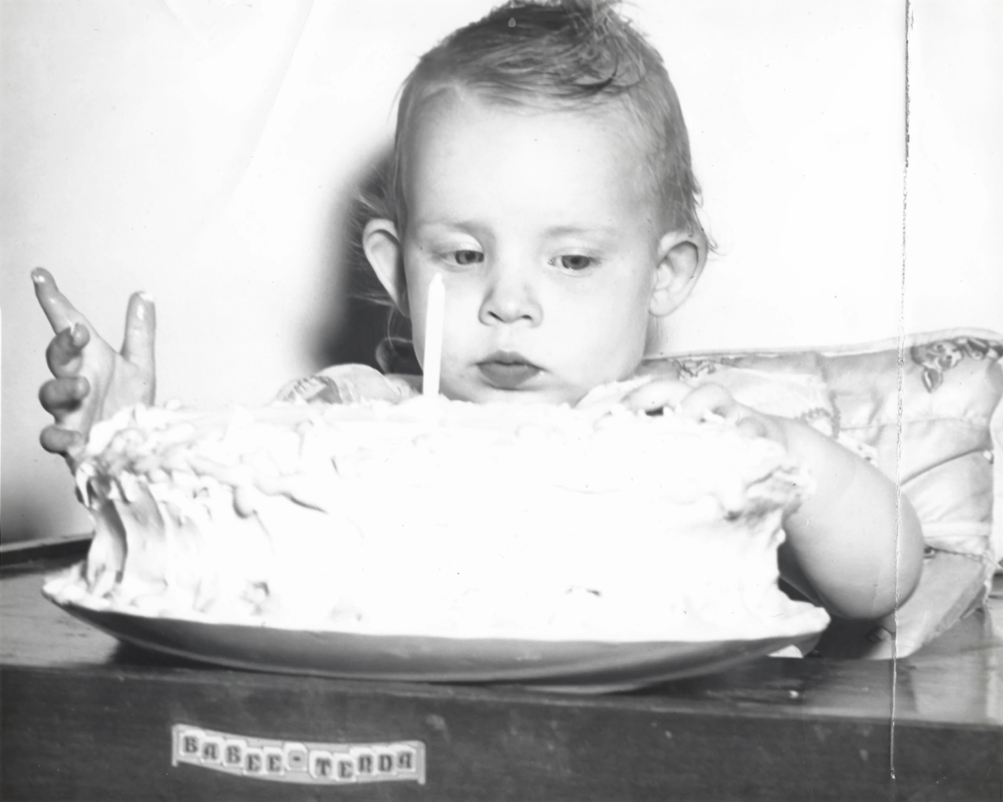 Cacky (b.1947) in a 'Babee Tenda' feeding table reaching toward a large white frosted first-birthday cake with a single lit candle, around 1948