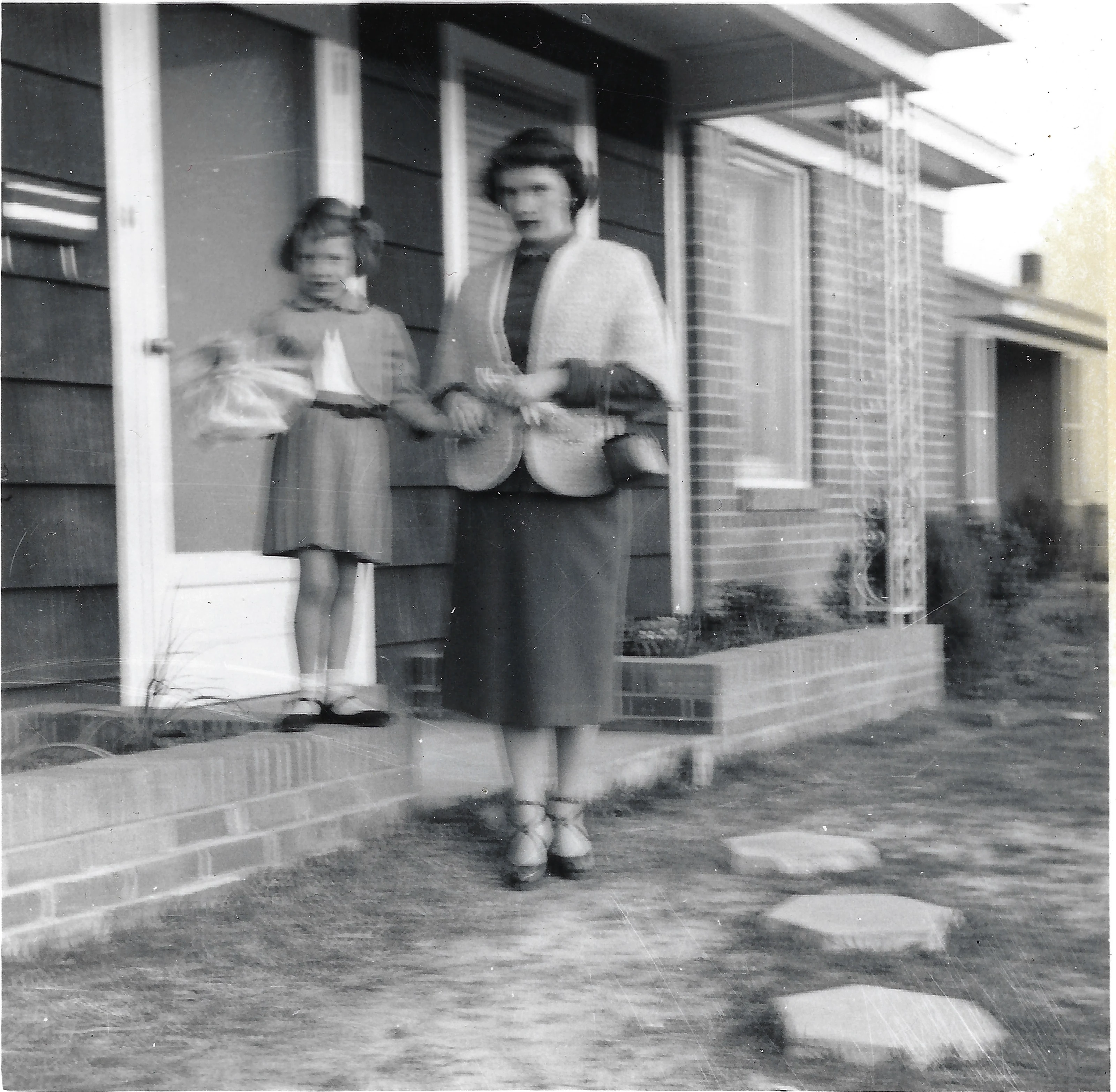 Cacky as a young girl holding a basket beside her mother, Gruzel, in a skirt suit and knit shawl on the front walk of a house, around 1955