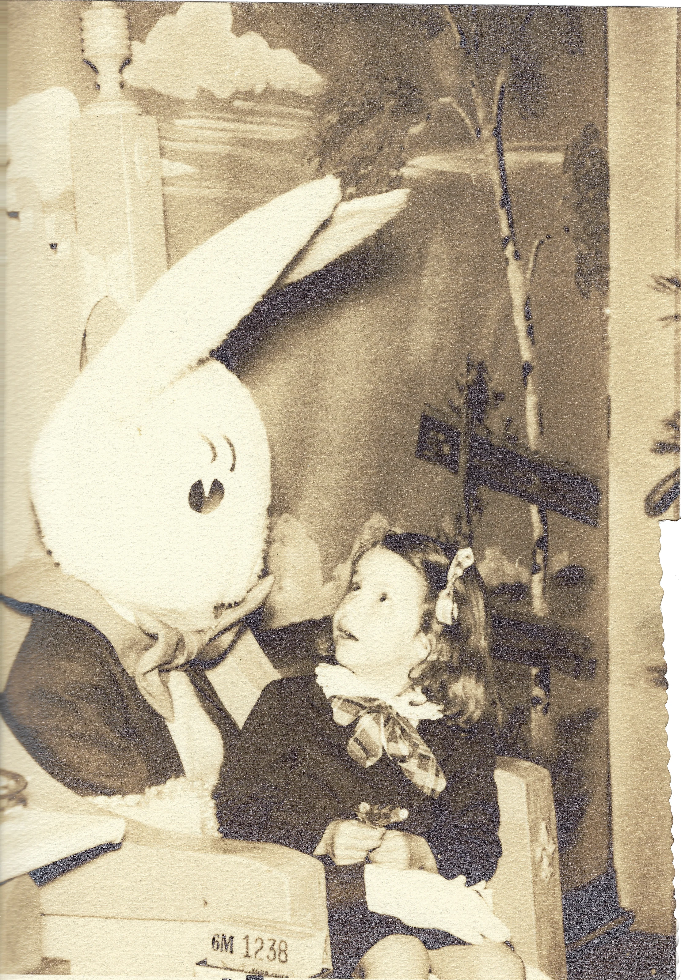 Cacky (b.1947) seated beside a person in an Easter Bunny costume before a decorated backdrop, sepia print, around 1955