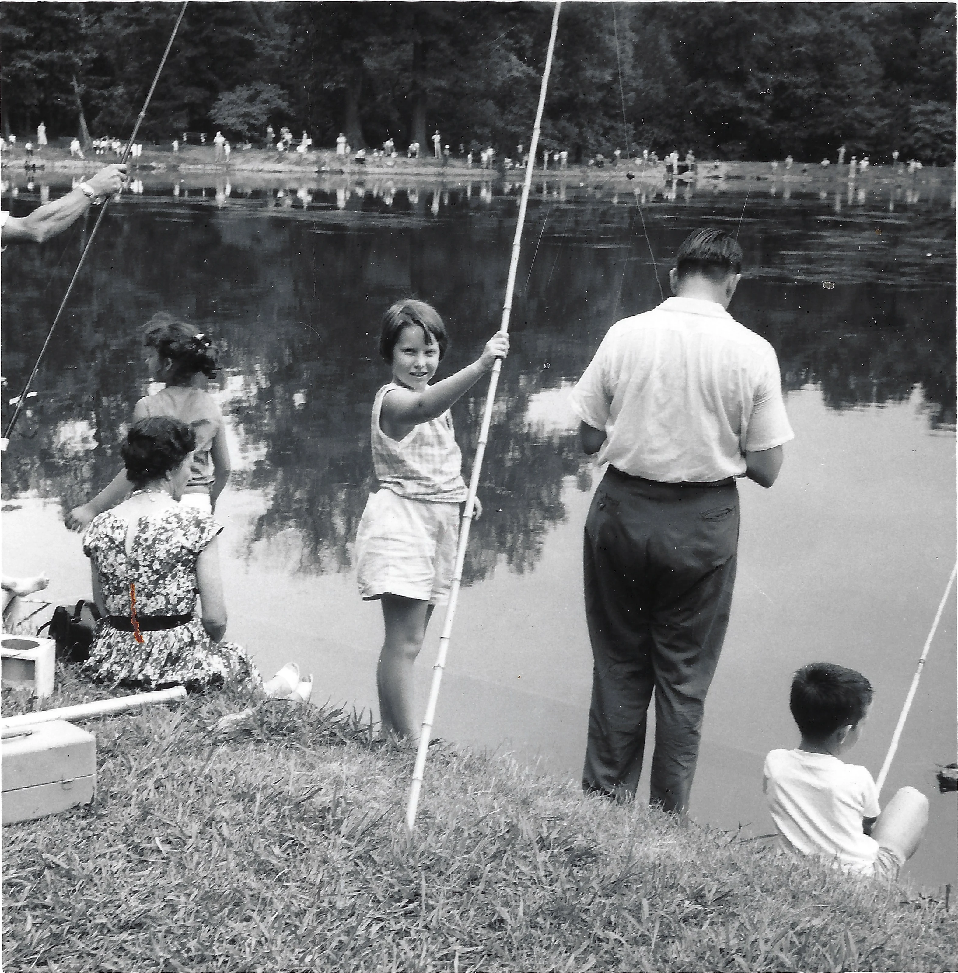 Cacky (b.1947) with a bamboo fishing pole smiling at the camera at a crowded fishing pond, with adults and another child nearby, around 1955