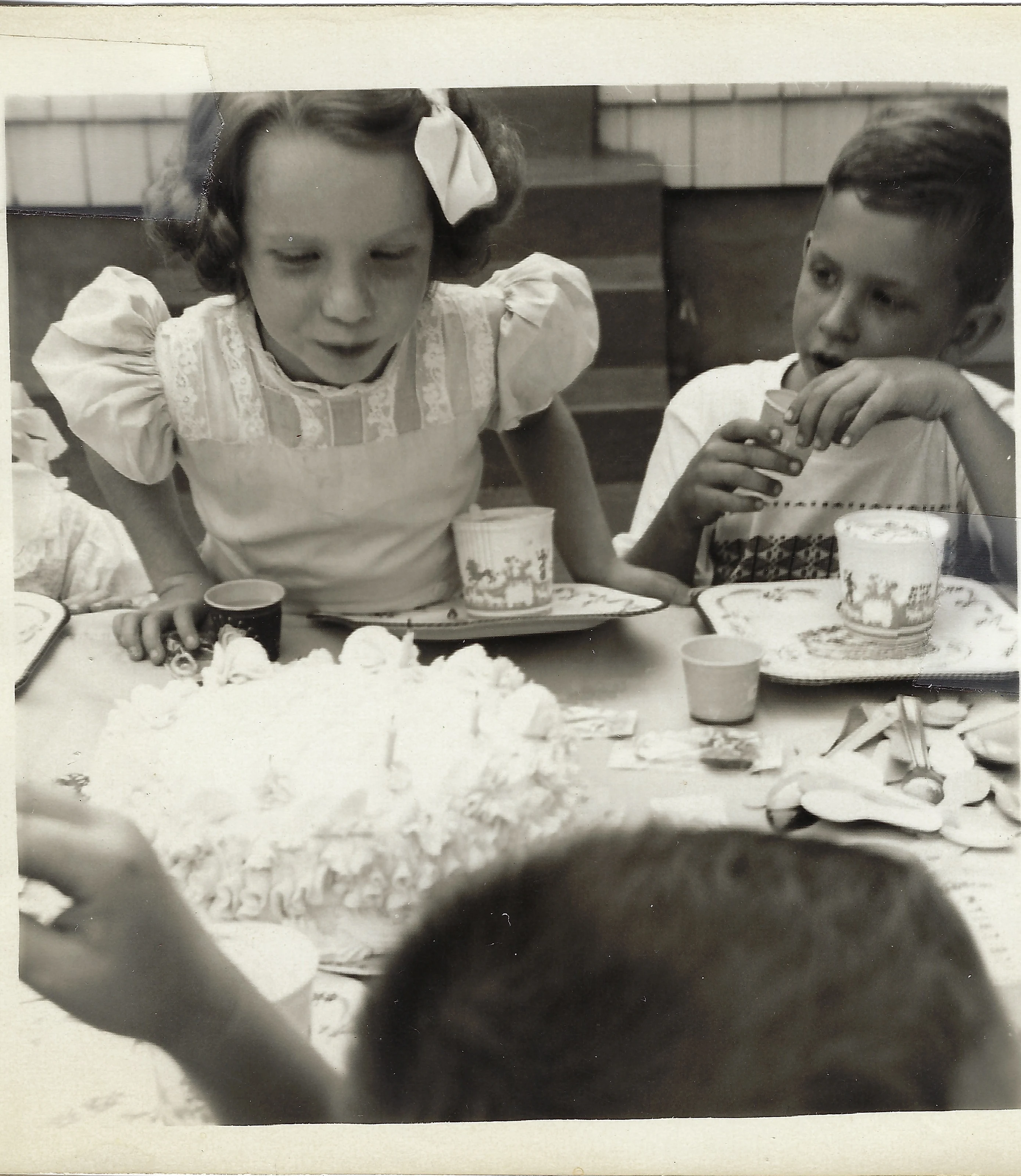 Cacky (b.1947) in a white party dress leaning over a large frosted cake at an outdoor birthday party while a boy eats nearby, around 1955