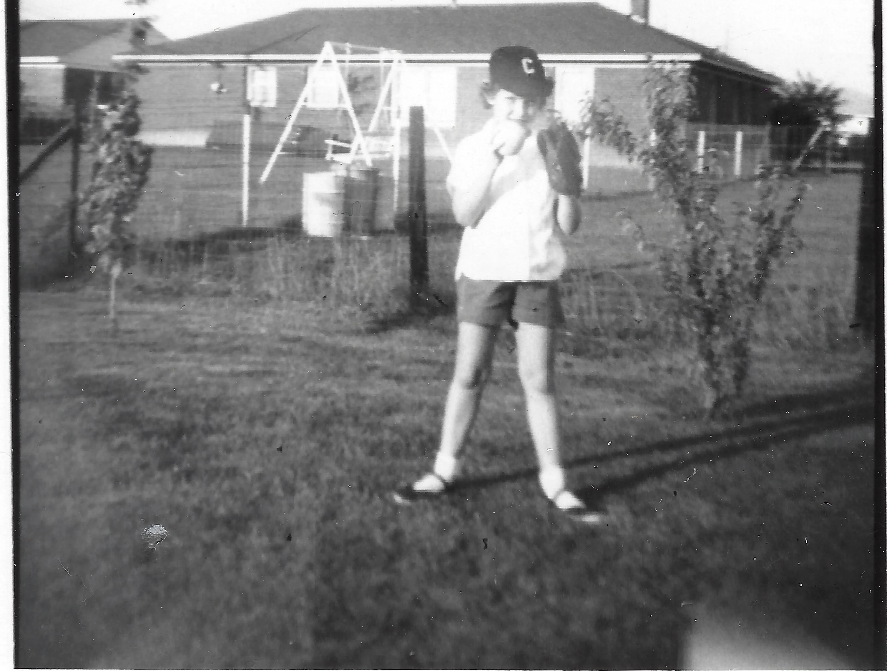 Cacky as a girl in a 'C' ball cap and shorts in a pitching stance in the backyard, a swing set behind, around 1956