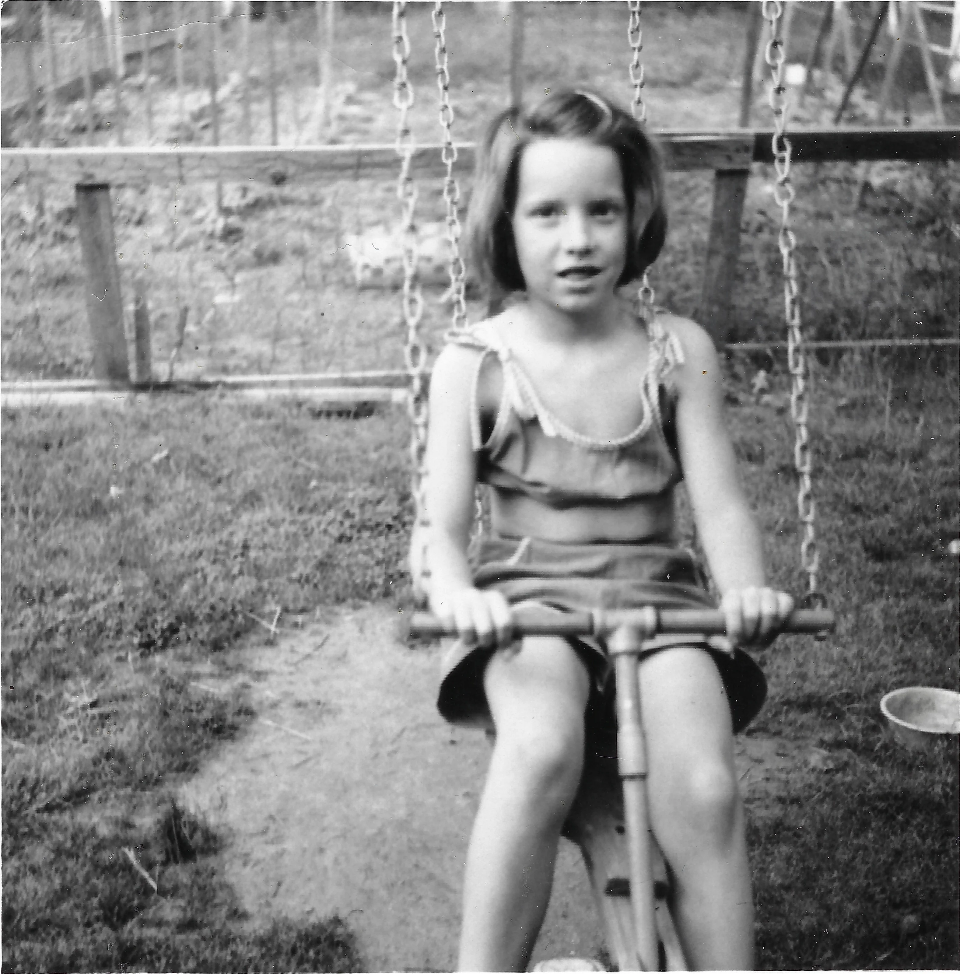 Cacky as a girl in a sunsuit on a chain swing in the backyard garden, around 1956
