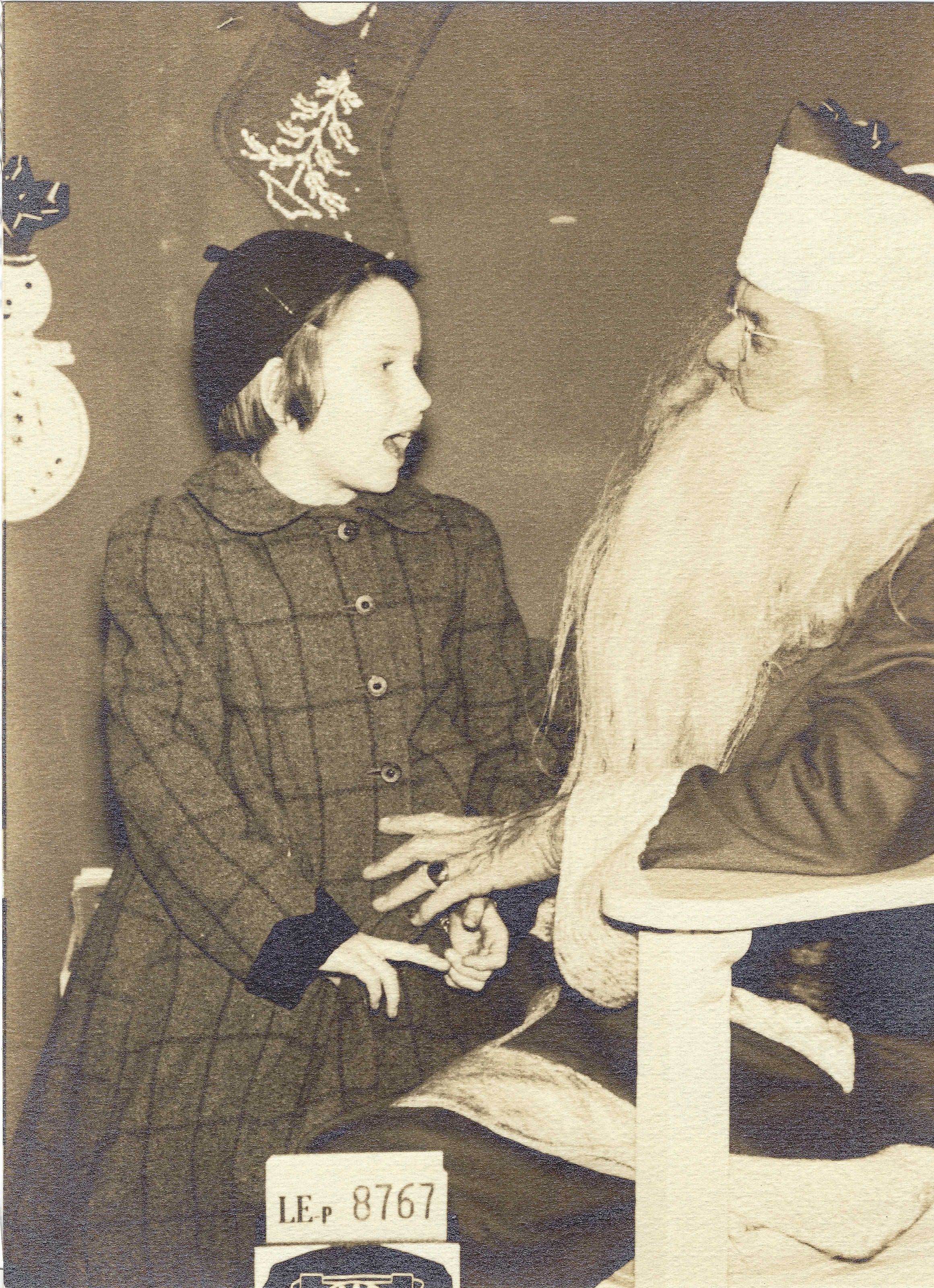 Cacky (b.1947) in a plaid coat and hat sitting with Santa, sepia studio print, around 1956