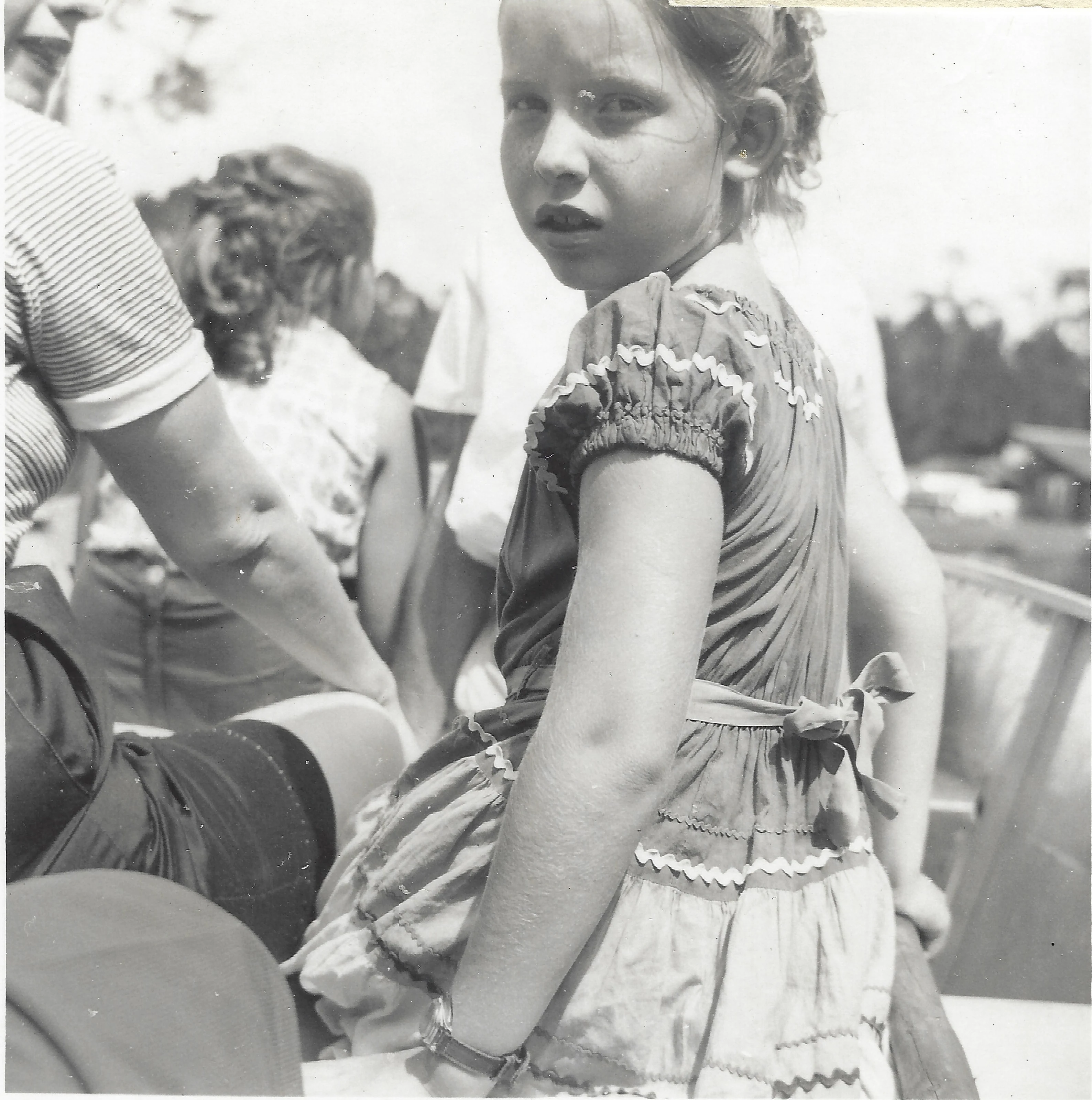Cacky (b.1947) in a ric-rac-trimmed fiesta dress looking over her shoulder at an outdoor gathering, around 1956