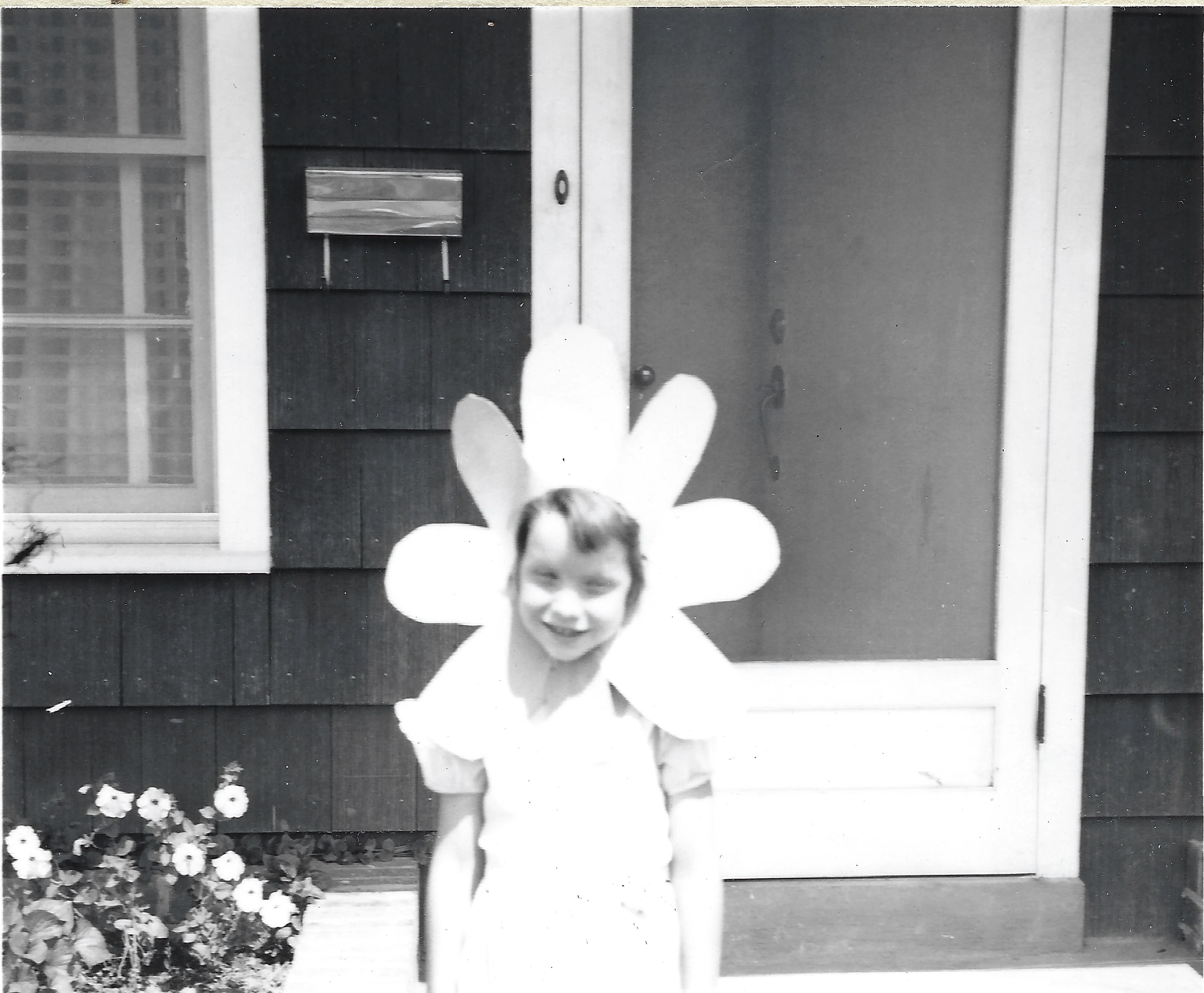 Cacky as a girl in a white dress and a large daisy-petal headpiece costume, standing before a house with flowers in the foreground, around 1956