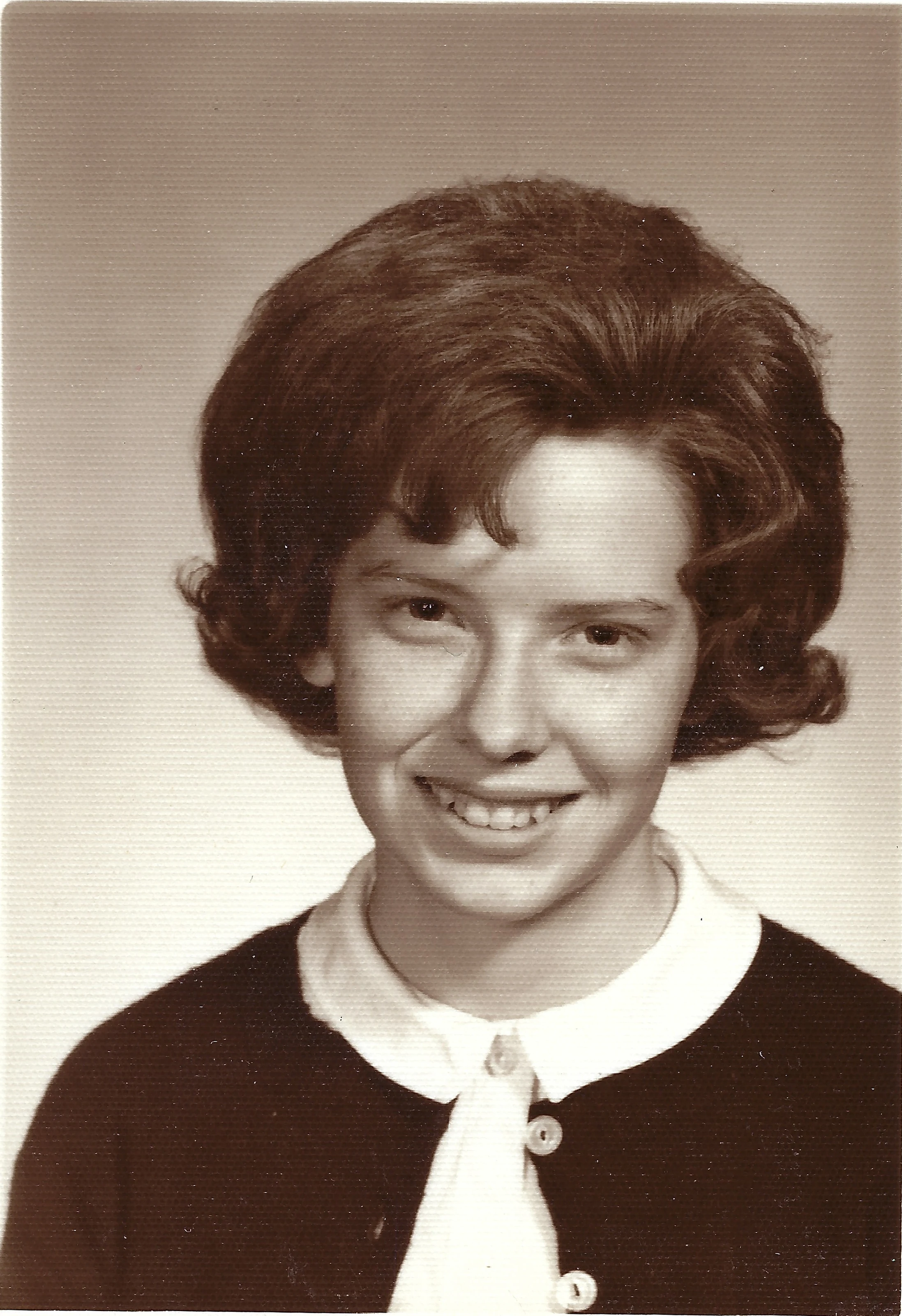 A school portrait of Cacky as a teenager in a dark cardigan over a white blouse with a tie at the collar, around 1962