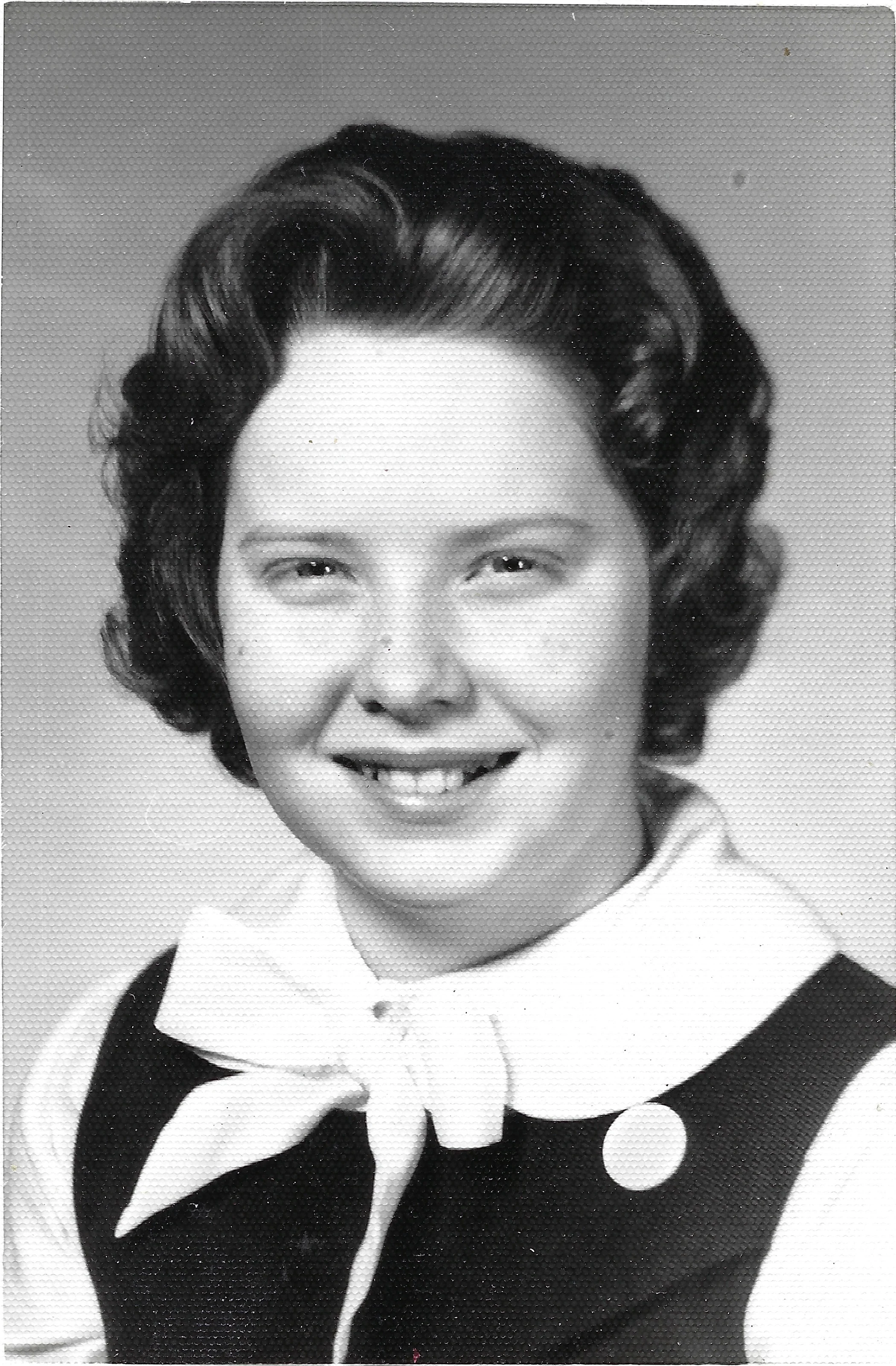 A black-and-white school portrait of Cacky as a teenager in a dark jumper, white blouse, and bow, around 1962