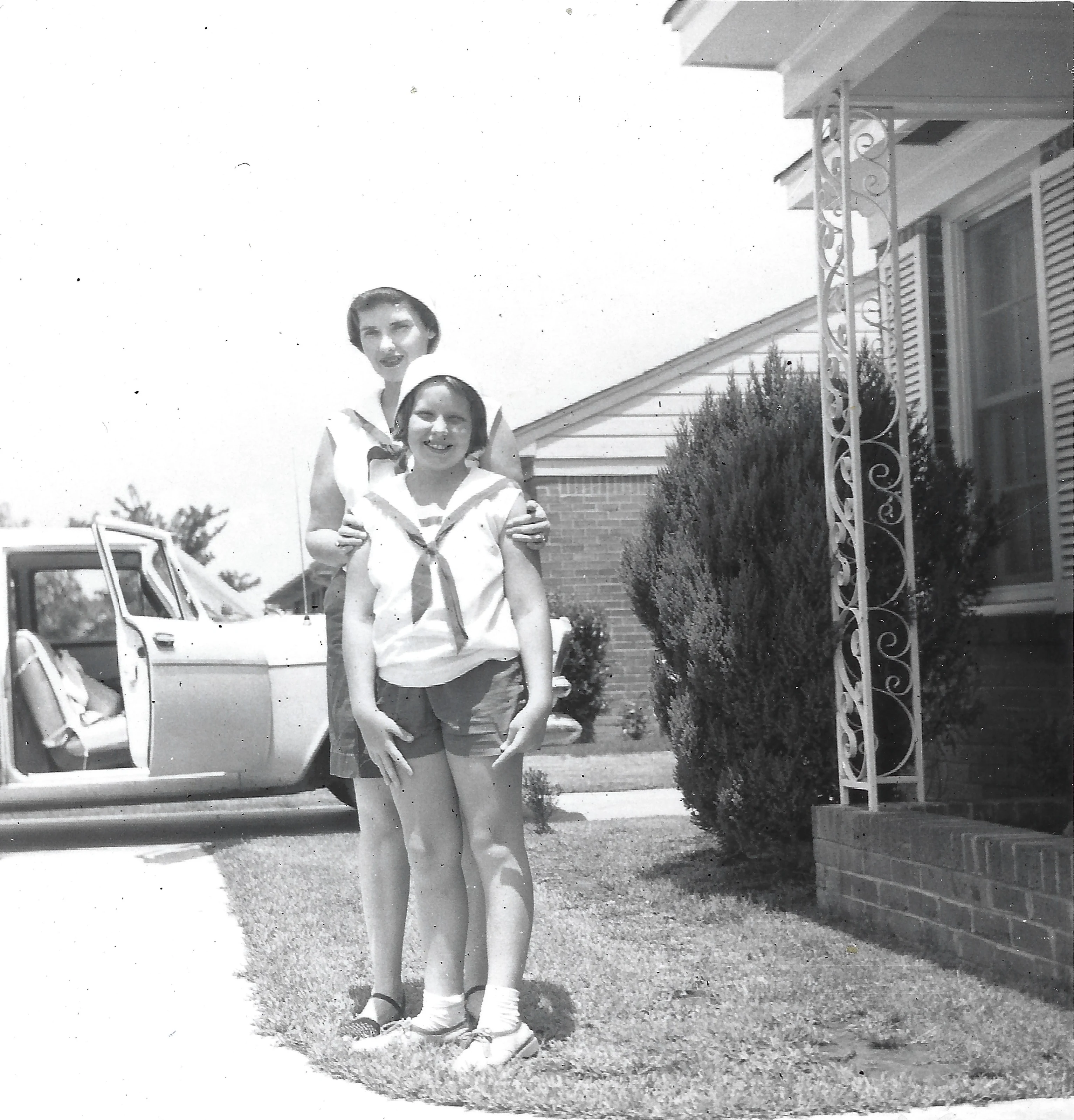 Gruzel (b.1924) with her hands on the shoulders of her daughter Cacky in a sailor top, in a front yard beside a car, around 1957