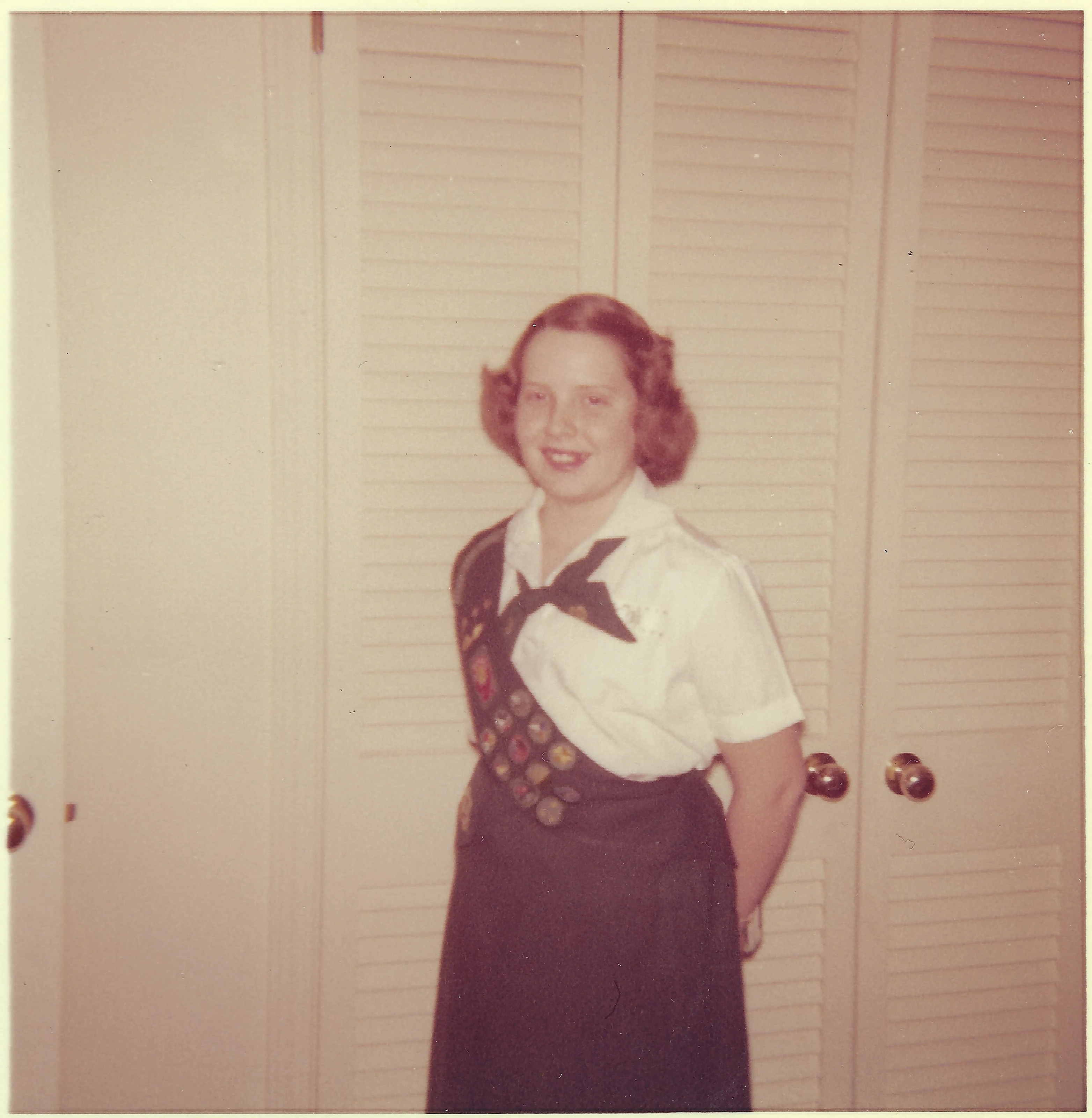 Cacky as a teenager in a Girl Scout uniform with a badge sash, standing by louvered doors, around 1962