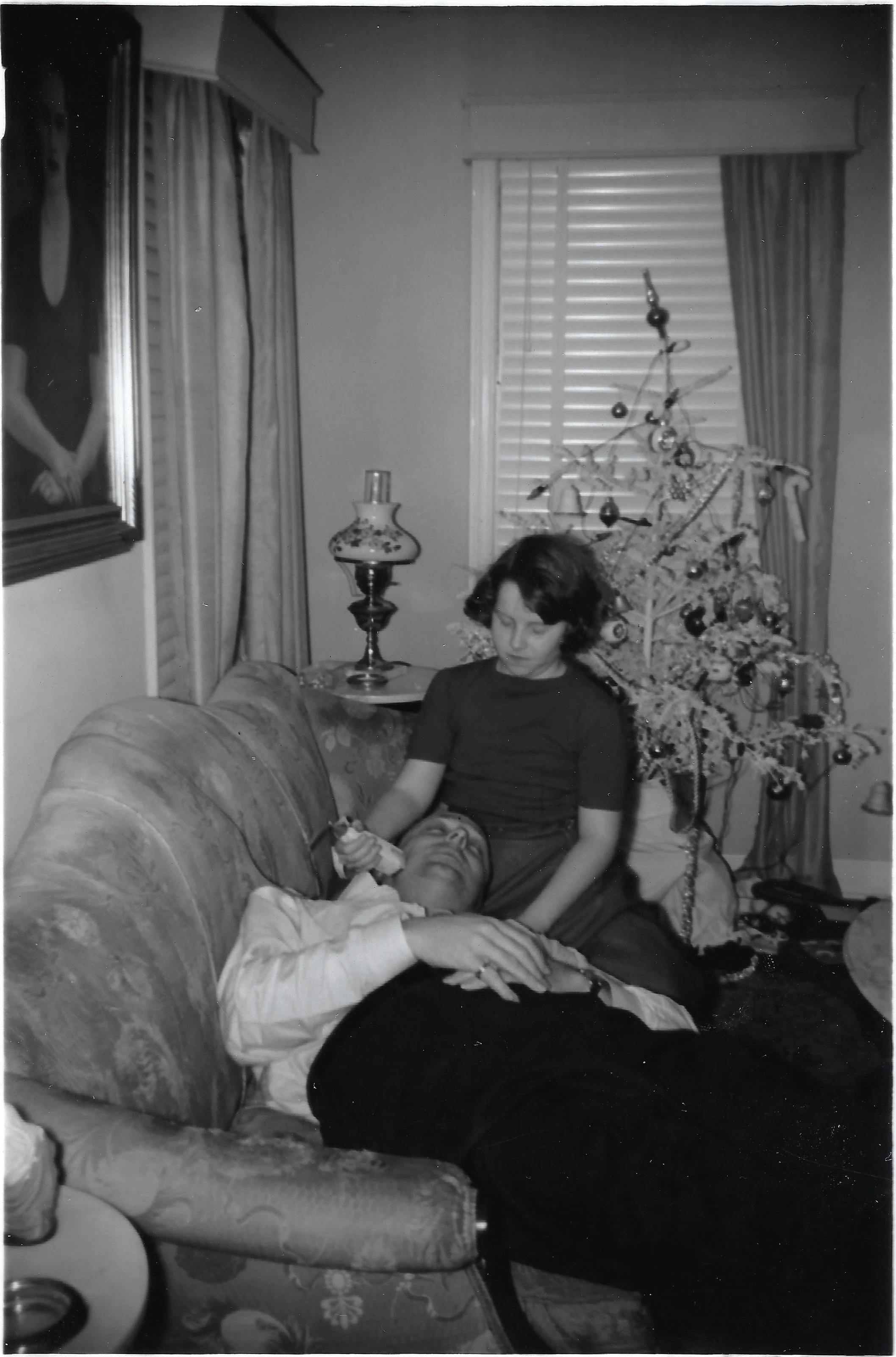 Cacky (b.1947) seated on a sofa cradling the head of her father, David 'Big Goose' Wilkins, who rests in her lap, beside a small decorated Christmas tree; a framed portrait of Gruzel hangs on the wall, around 1964