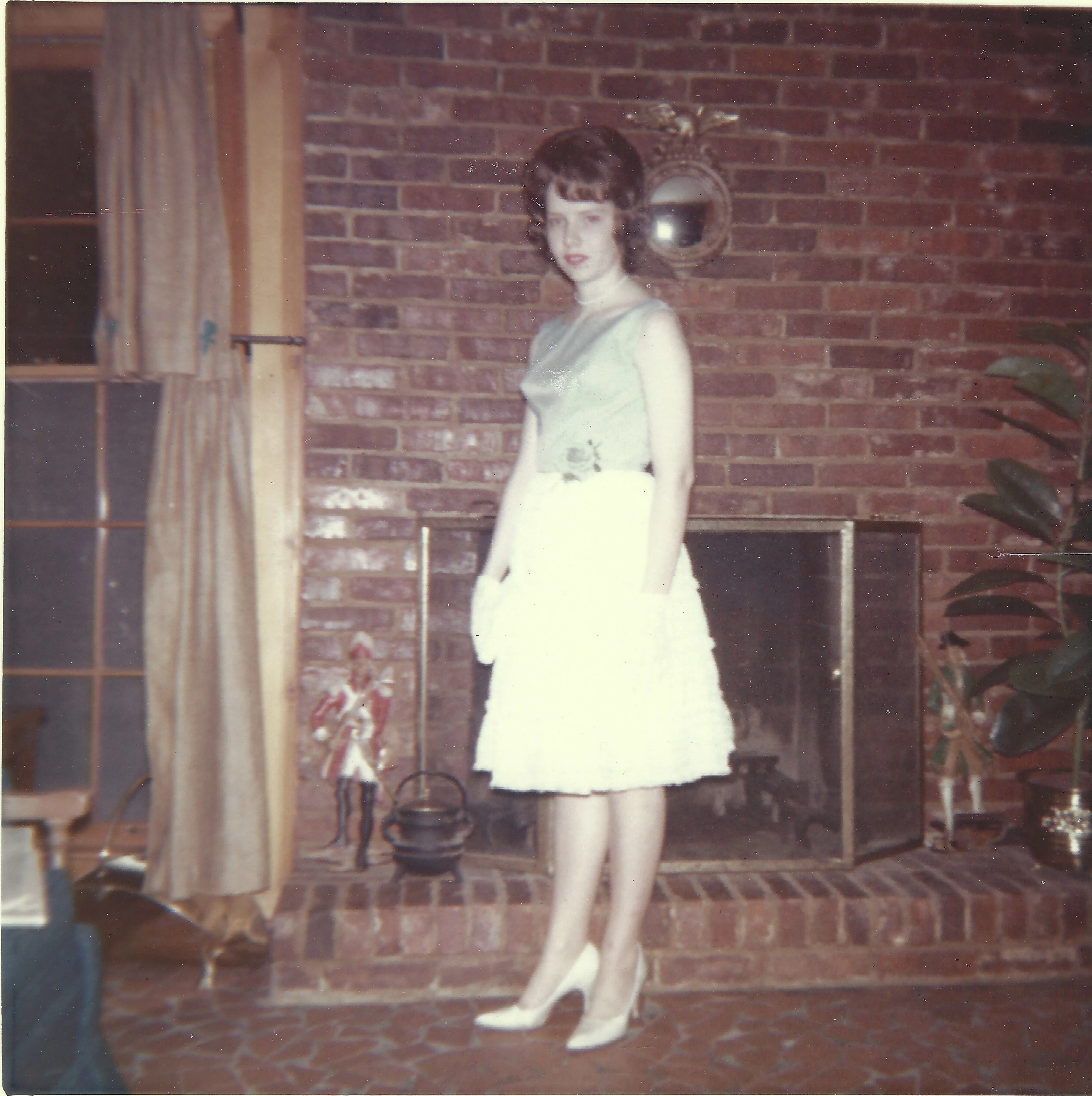 Cacky as a young woman in a green top, white skirt, and heels standing by a brick fireplace, around 1966