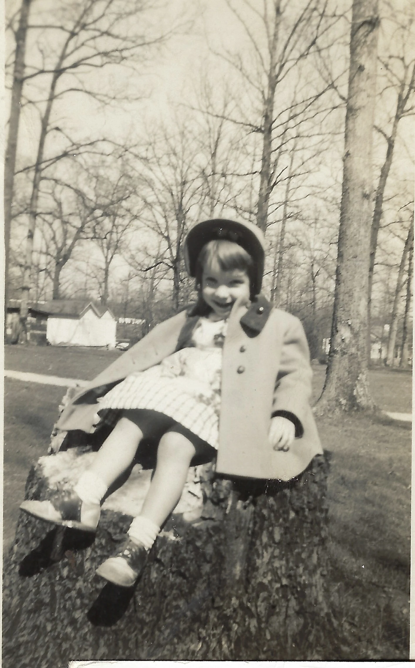Cacky as a young girl in a plaid dress with a coat over her shoulders and a hat, seated on a large tree stump among bare winter trees, around 1951