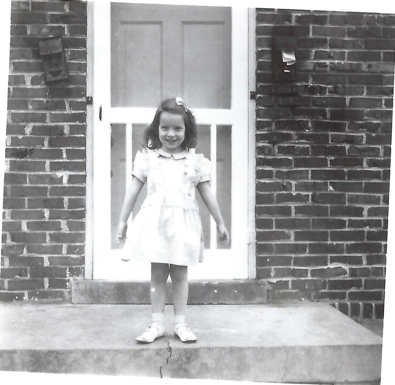 Cacky as a young girl with a hair bow, in a light party dress and white shoes, standing on a concrete stoop before the screen door of a brick house, around 1951