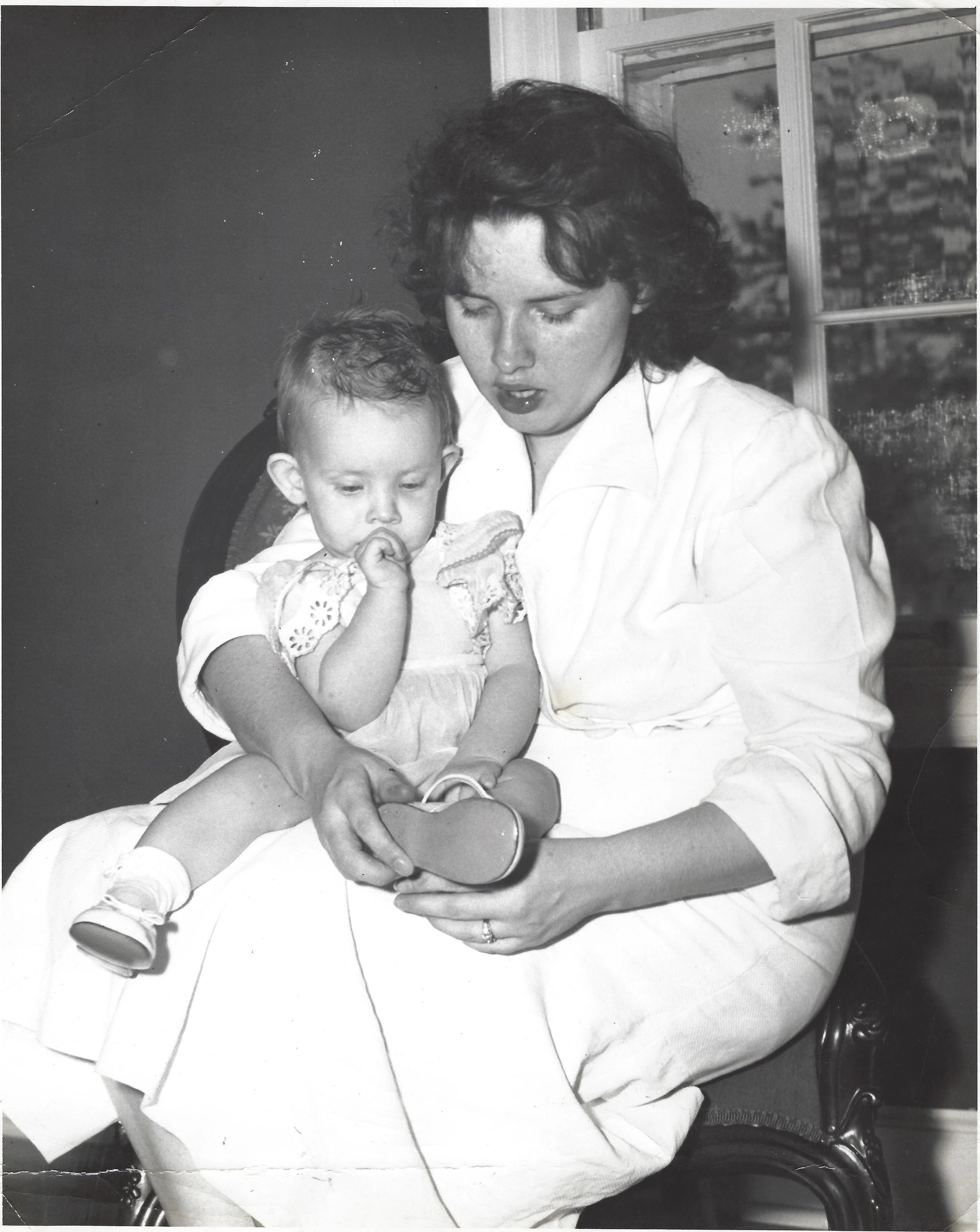 Gruzel (b.1924) in a white dress holding her baby daughter Cacky on her lap and fitting a small shoe, beside a window, around 1948