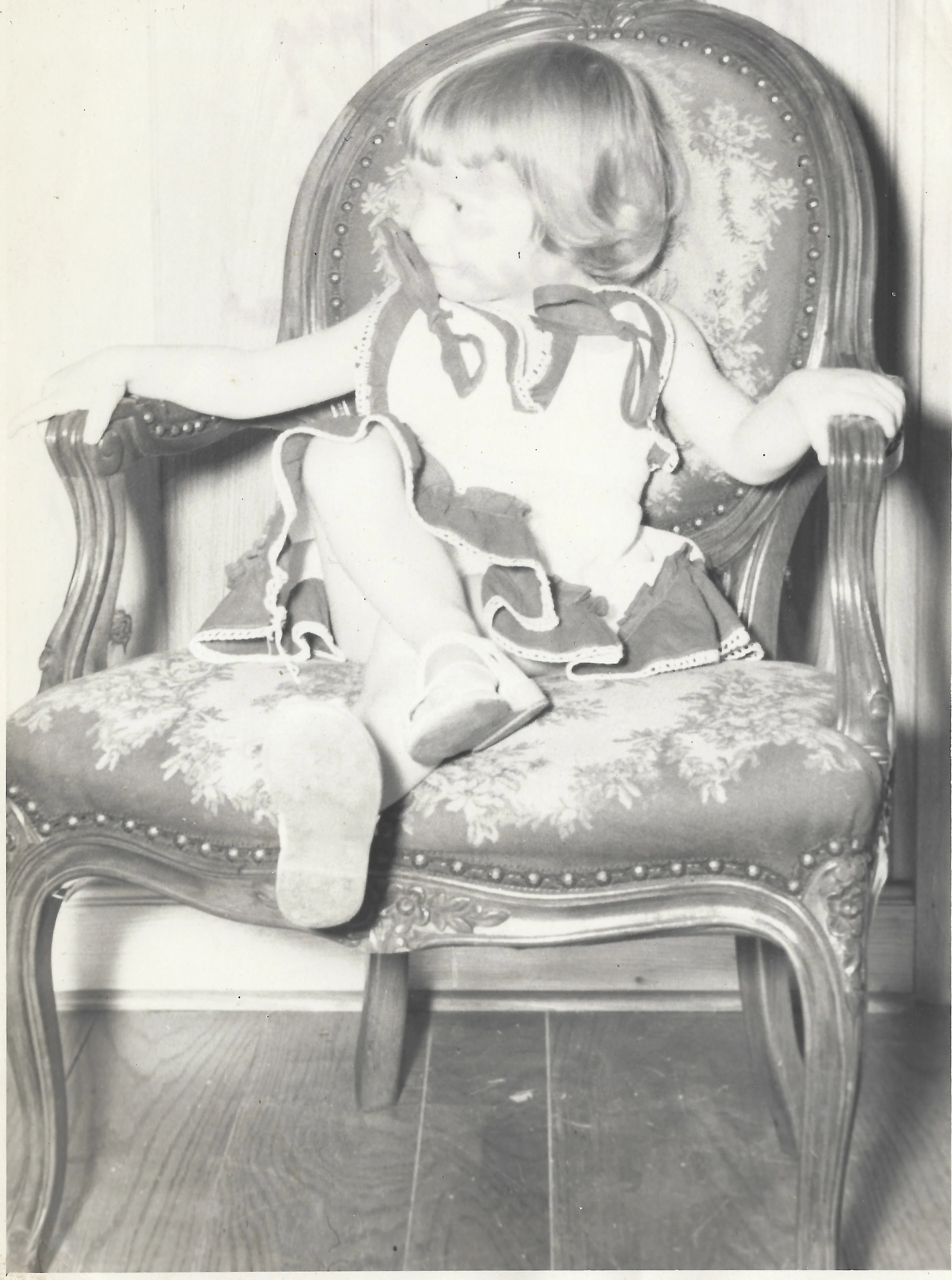 Cacky (b.1947) as a toddler in a ruffled pinafore and white socks, kneeling on a floral upholstered armchair, around 1950
