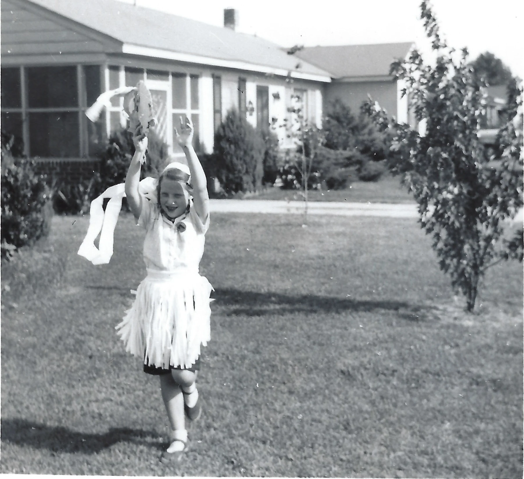 Cacky as a young girl in a fringed costume running across a front lawn with her arms raised, suburban houses behind, around 1953