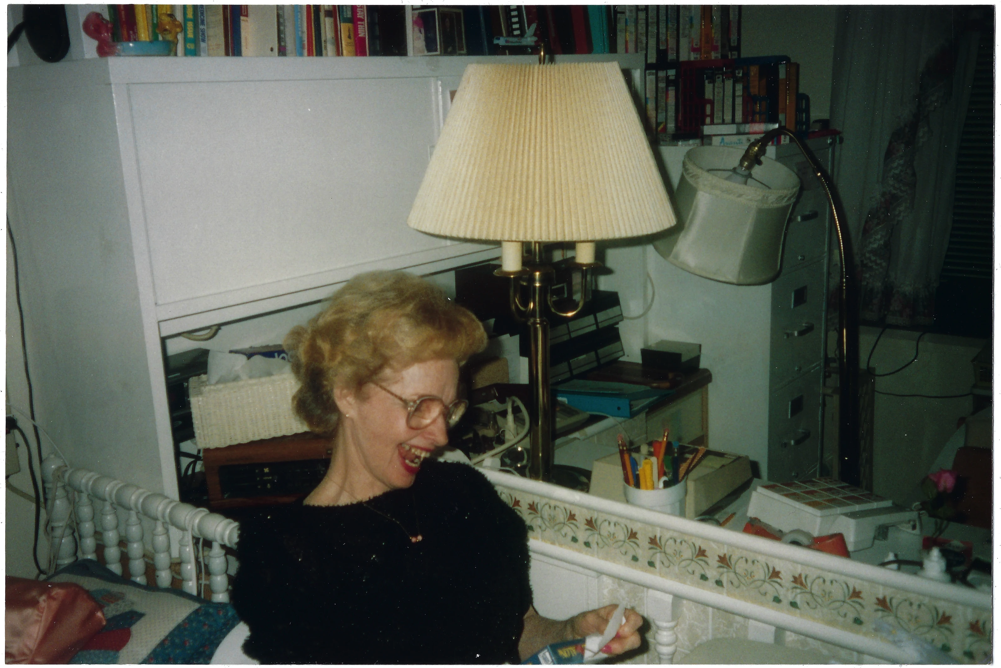 Gruzel (b.1924) laughing as she opens a new copy of the film 'Home Alone' in a book-filled room, around 1991