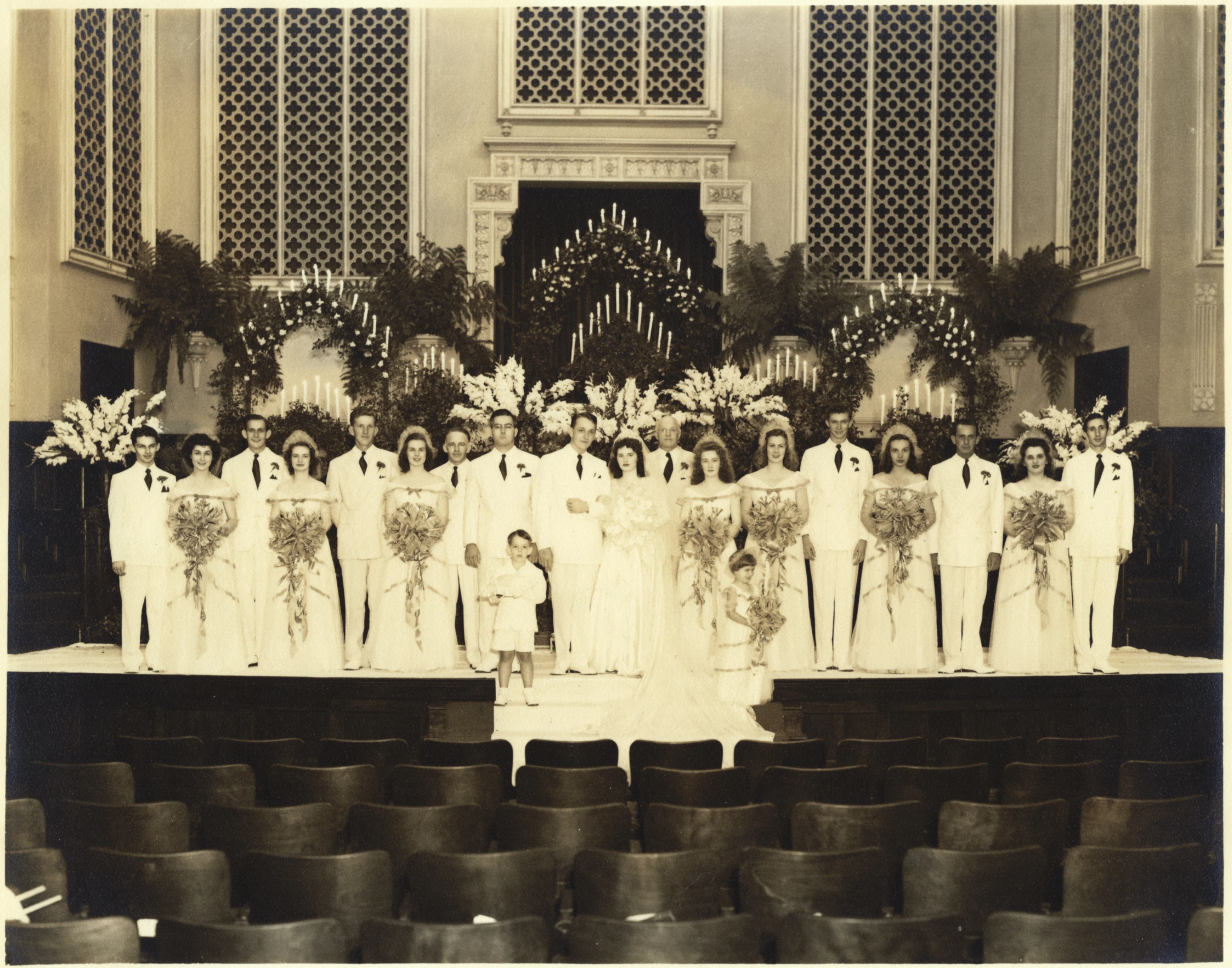 A large formal wedding party posed before flowers and candles in a church sanctuary, with two children in front, around 1946.
