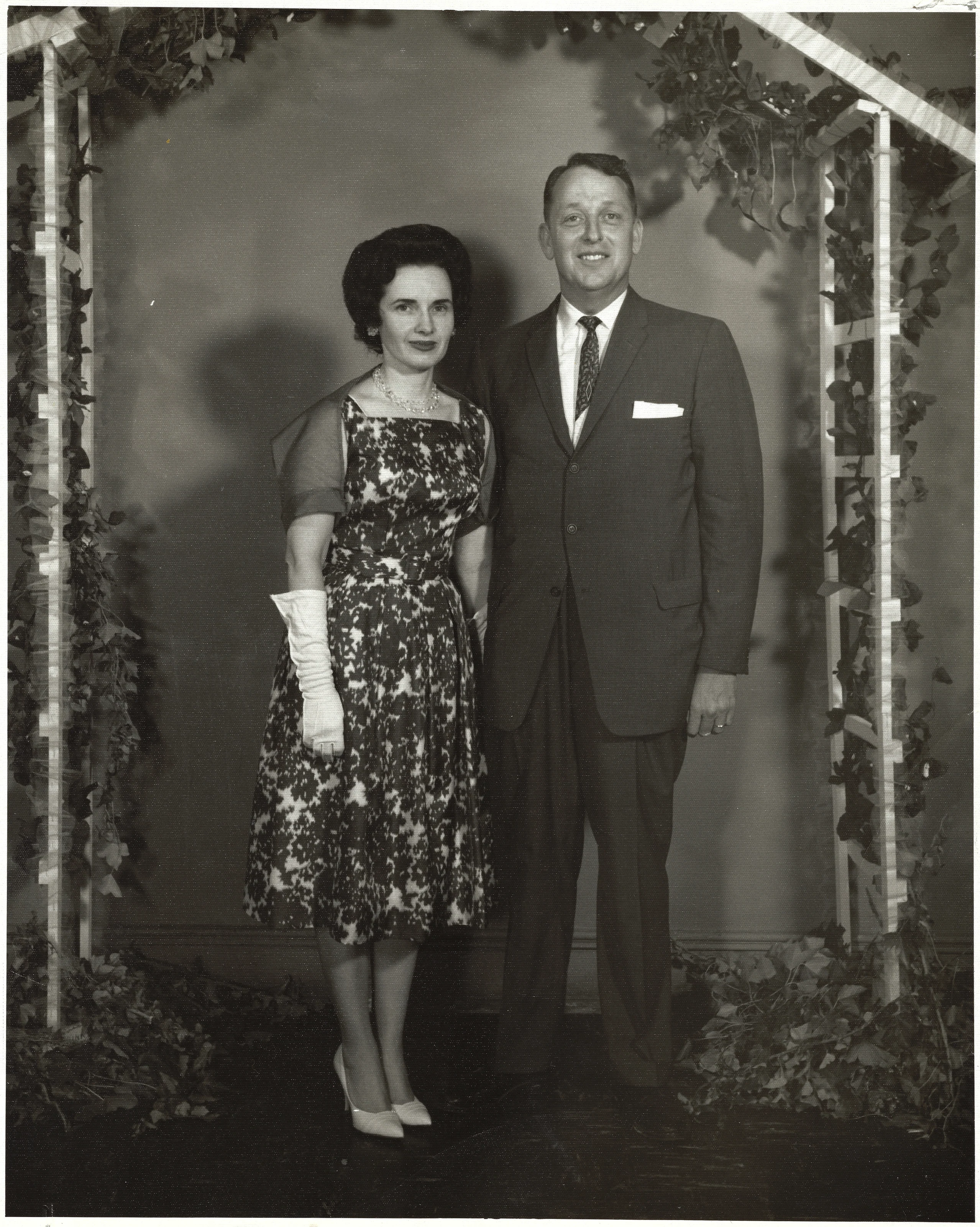 Gruzel (b.1924) in a floral dress and white gloves posed with her first husband, David Winborn 'Big Goose' Wilkins, under an ivy arch, around 1960