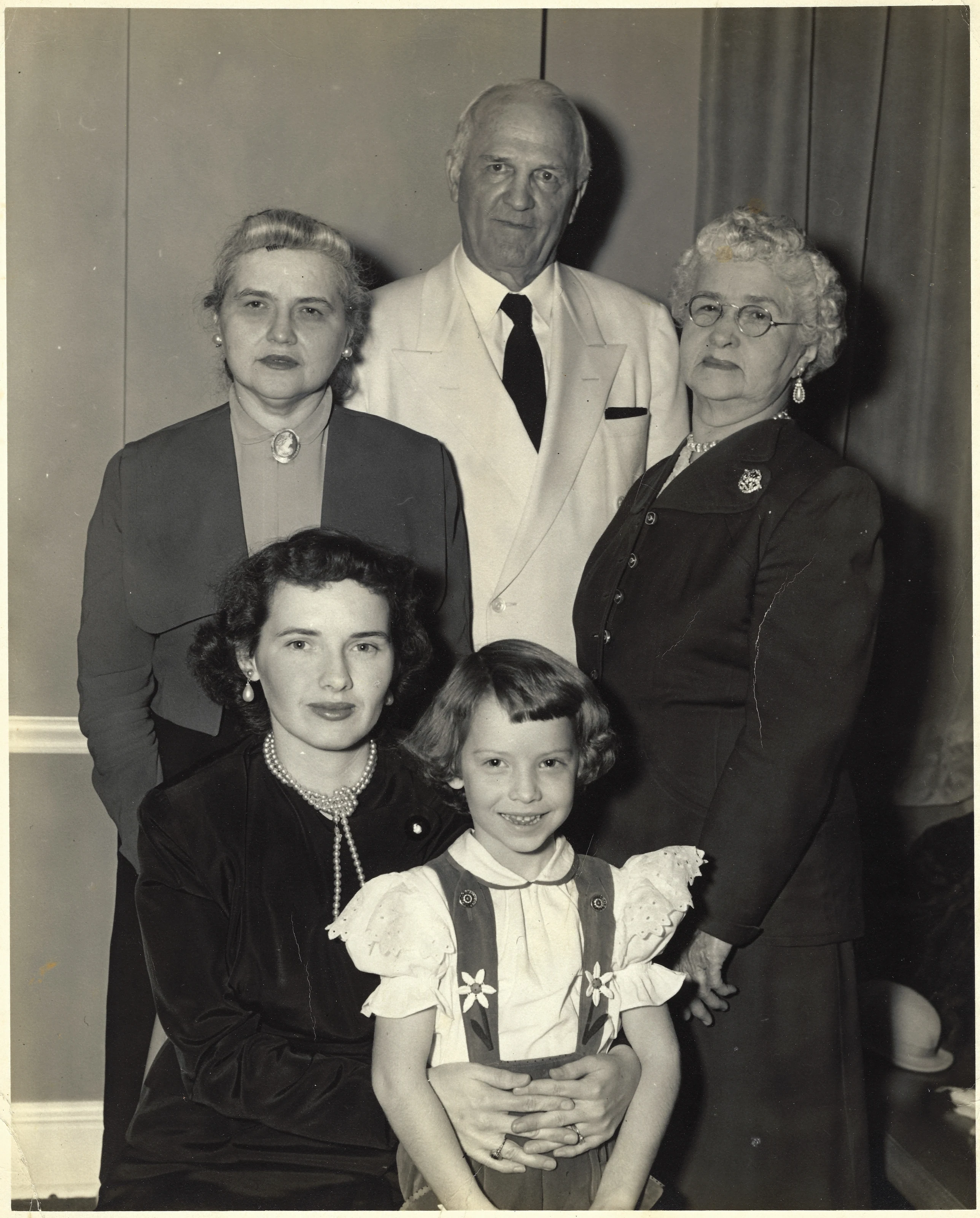 A mid-1950s family studio portrait: standing are Mama Percy (left) and her mother Alconia (right) with a gentleman between them; seated in front are Gruzel and her daughter Cacky (b.1947) as a young girl — four generations of the maternal line, around 1954