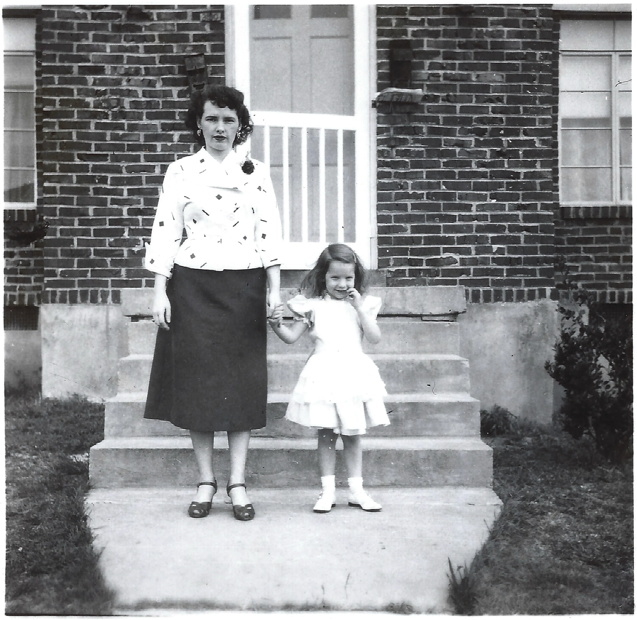 Gruzel (b.1924) in a patterned jacket holding hands with her daughter Cacky in a frilly white dress on the front steps of a brick house, around 1954