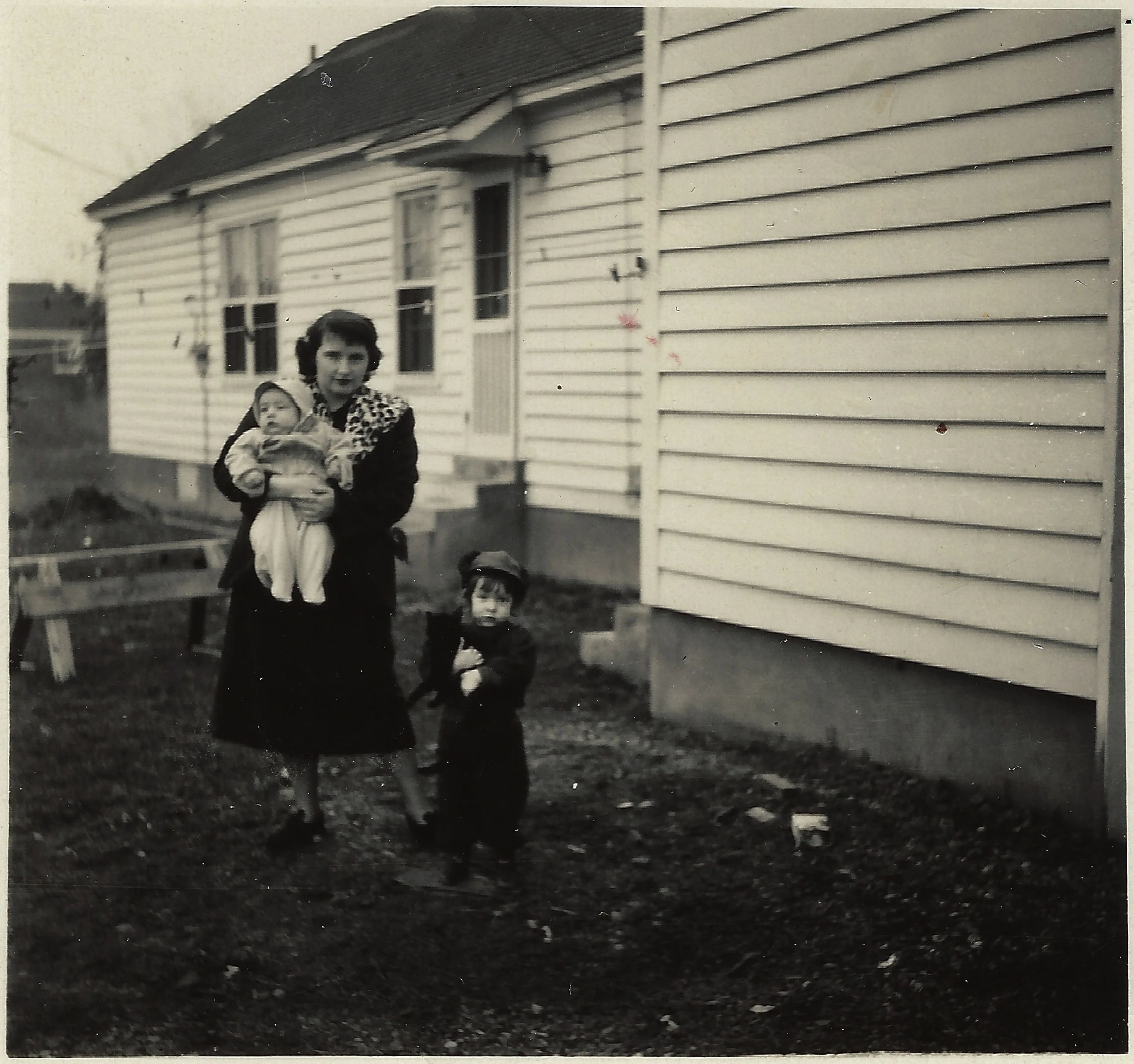 Gruzel (b.1924) in a dark leopard-collared coat holding a baby, a toddler in a cap beside her, in the yard by a clapboard house, around 1950