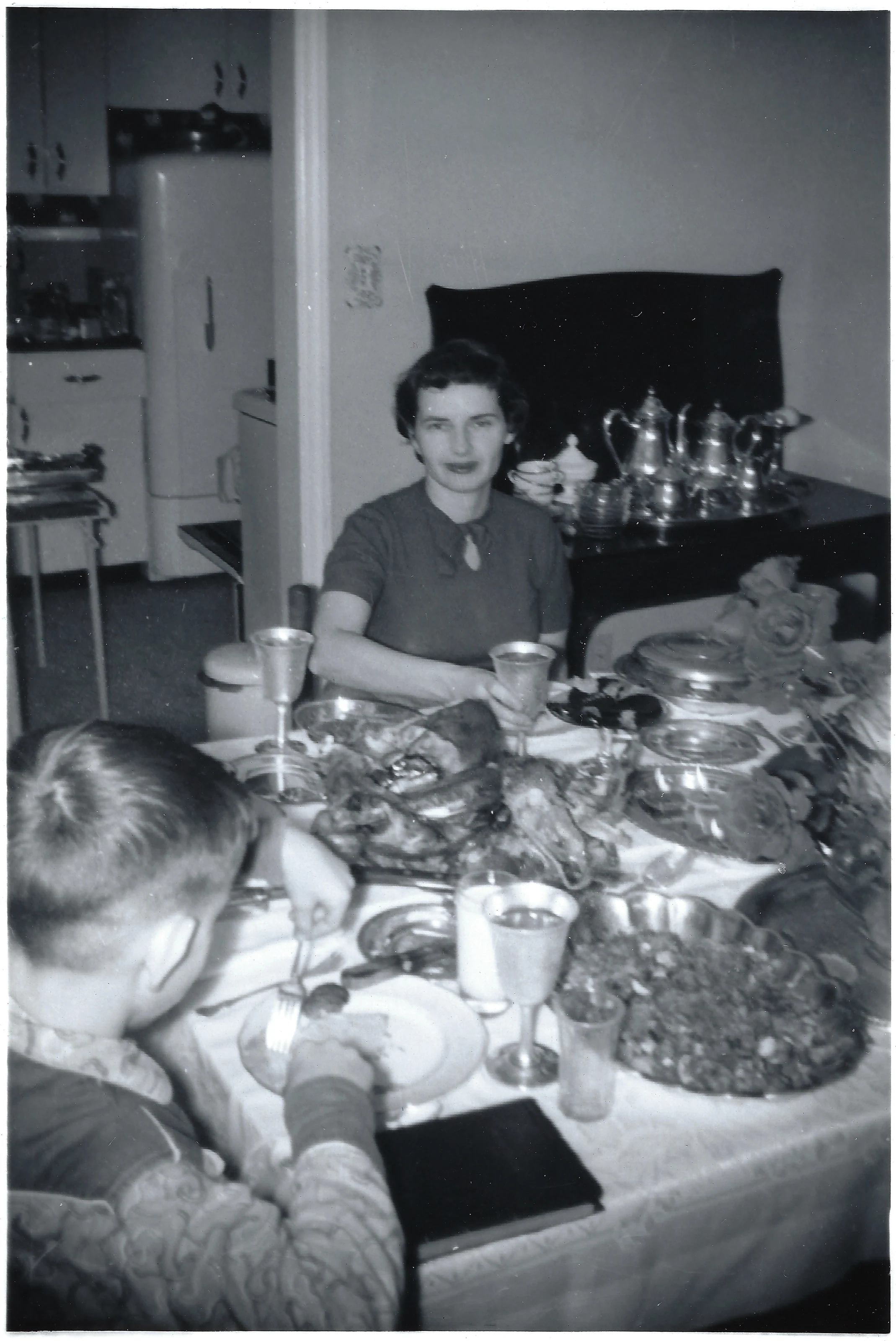 Gruzel (b.1924) seated at a laden holiday table with a silver coffee service, a boy eating in the foreground, around 1954
