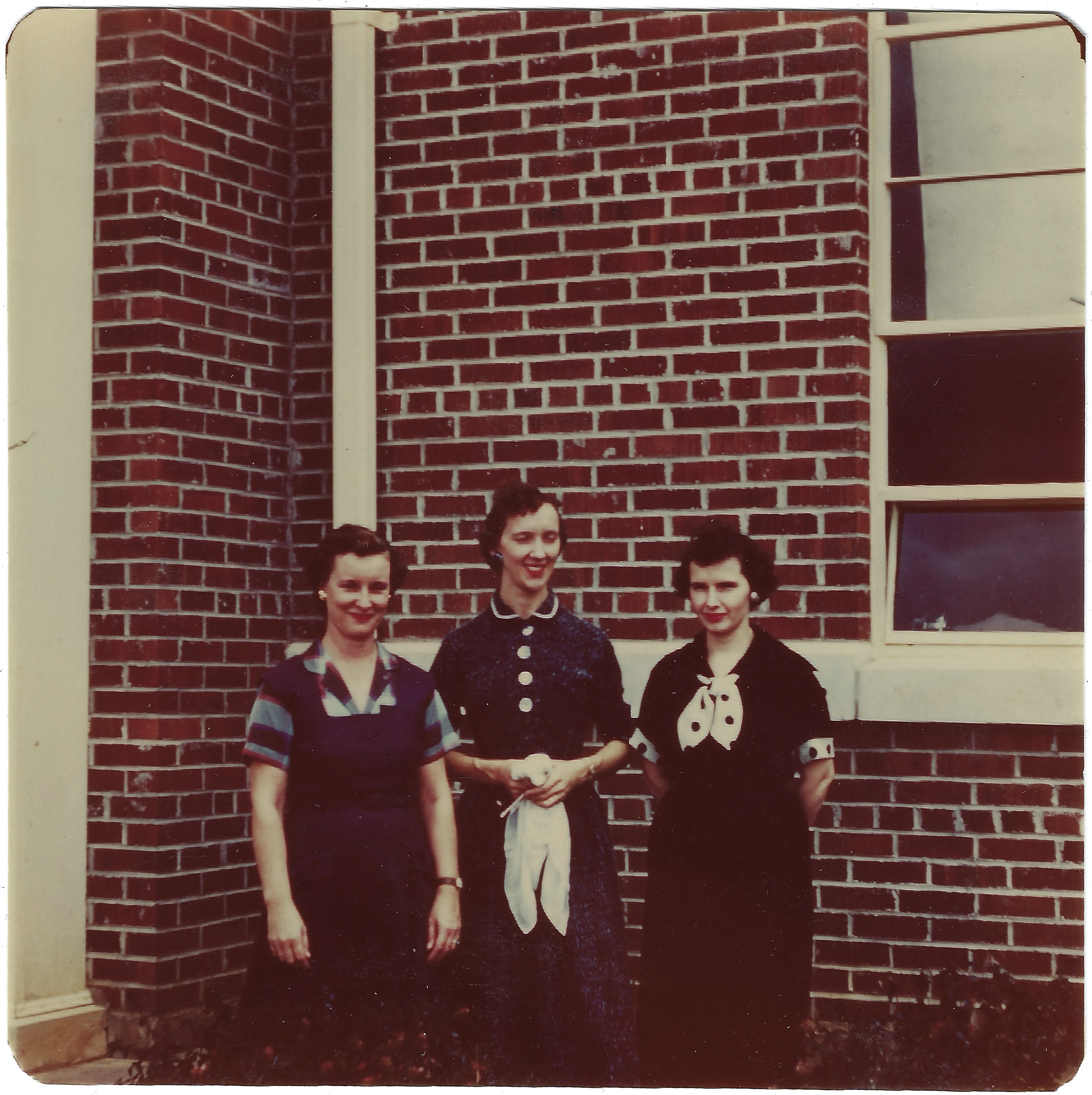 Three women in 1950s dresses standing together against a brick wall, Gruzel (b.1924) among them, around 1954