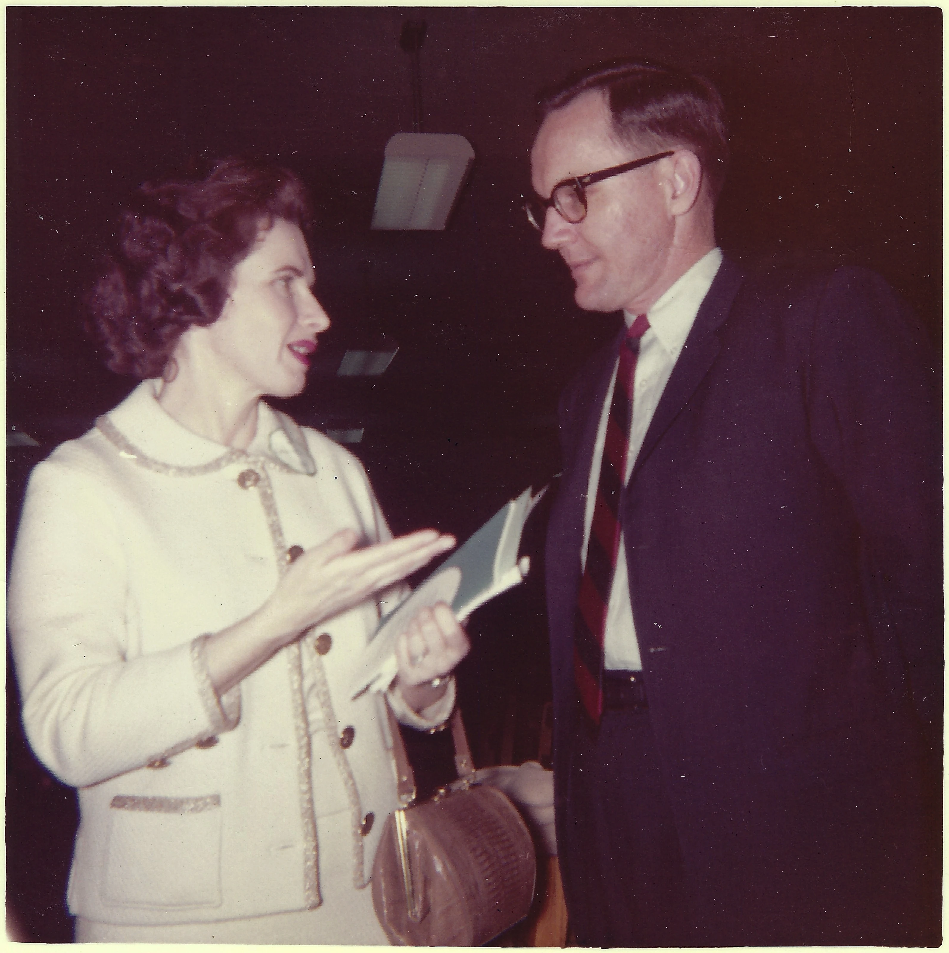 Gruzel (b.1924) in a white suit gesturing as she talks with a bespectacled man at an event, around 1962