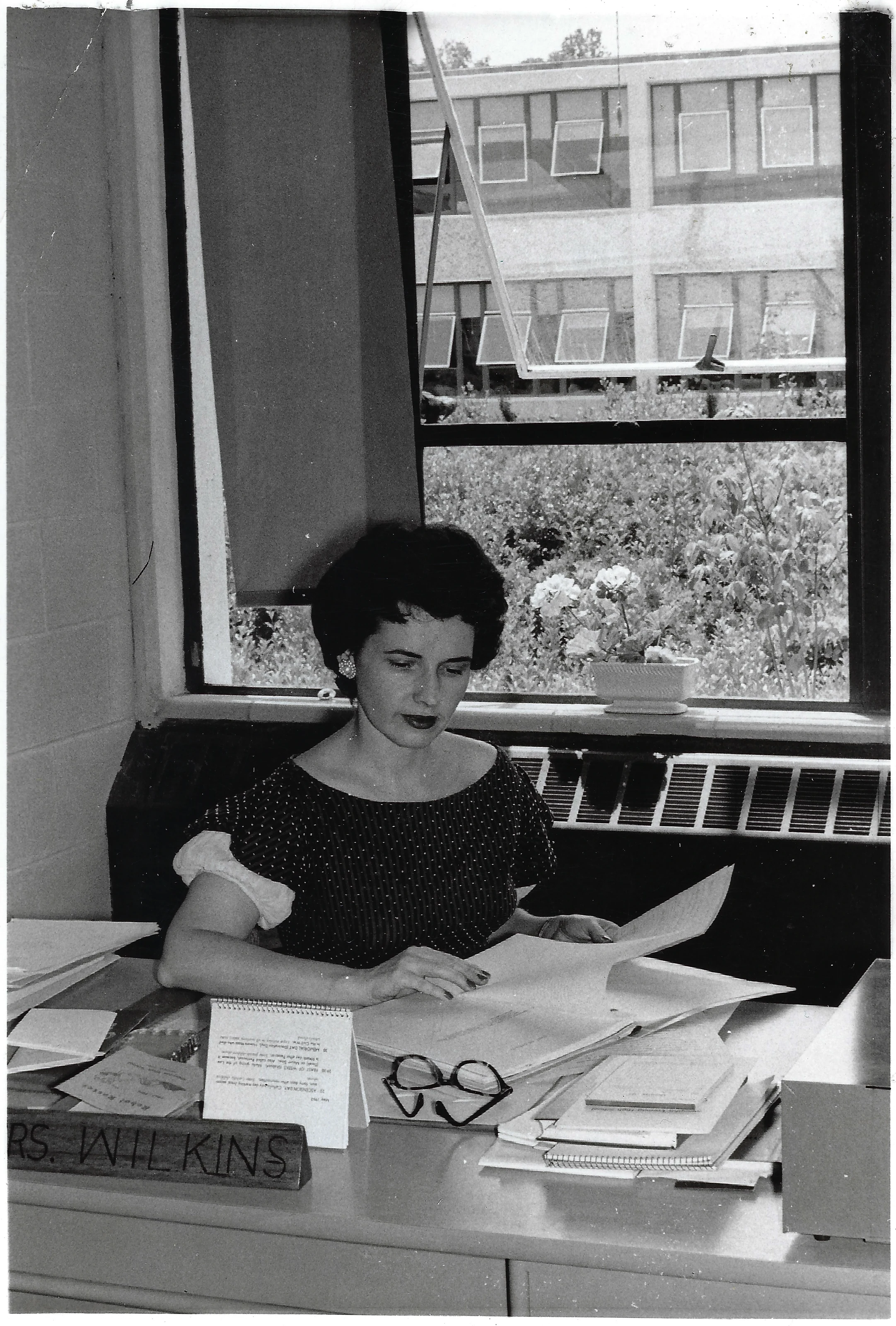 Gruzel (b.1924) working at a desk piled with papers by a window, a nameplate on the desk, around 1962