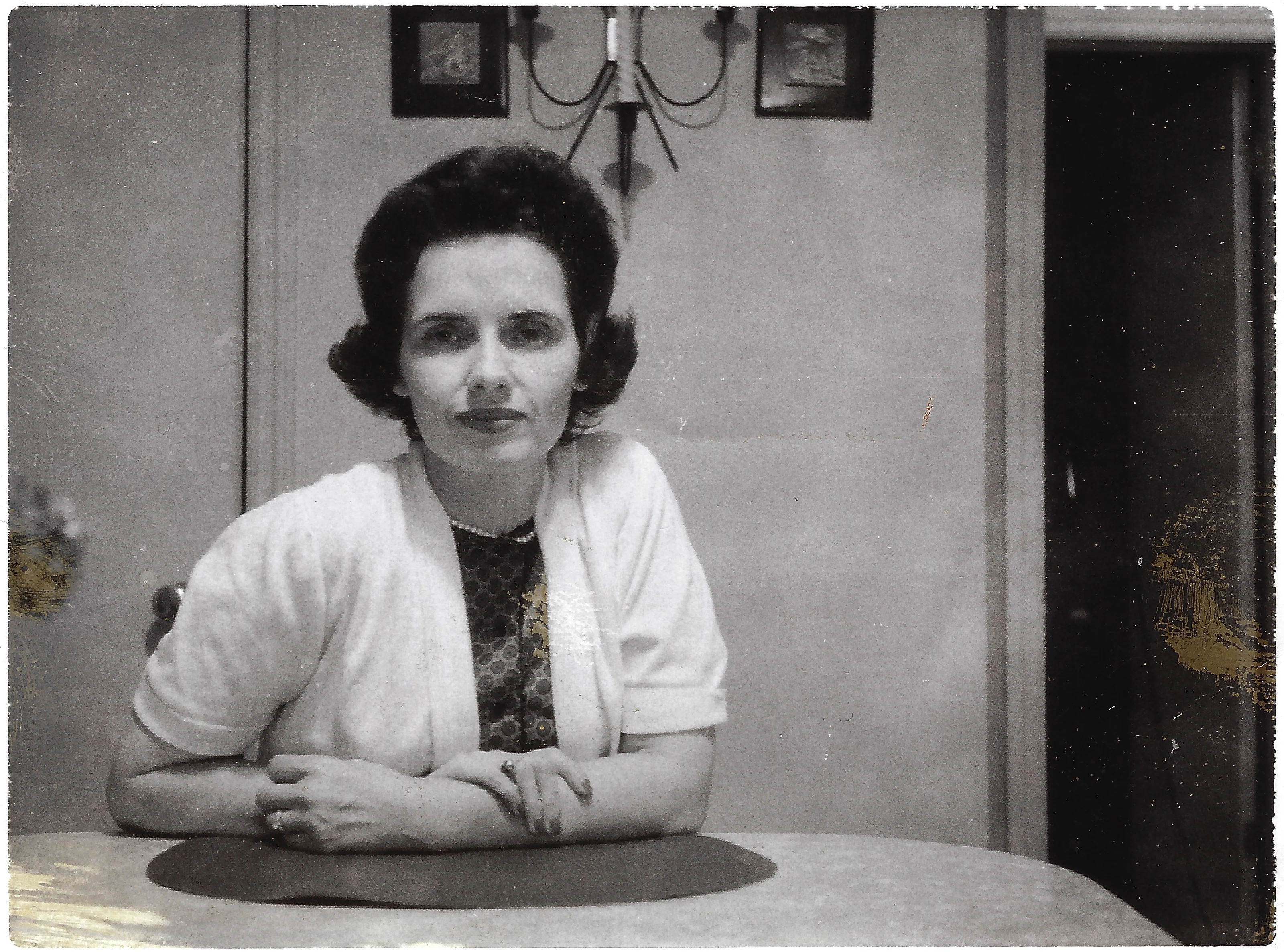A posed portrait of Gruzel (b.1924) seated at a table with her arms folded, in a cardigan over a print dress, around 1962