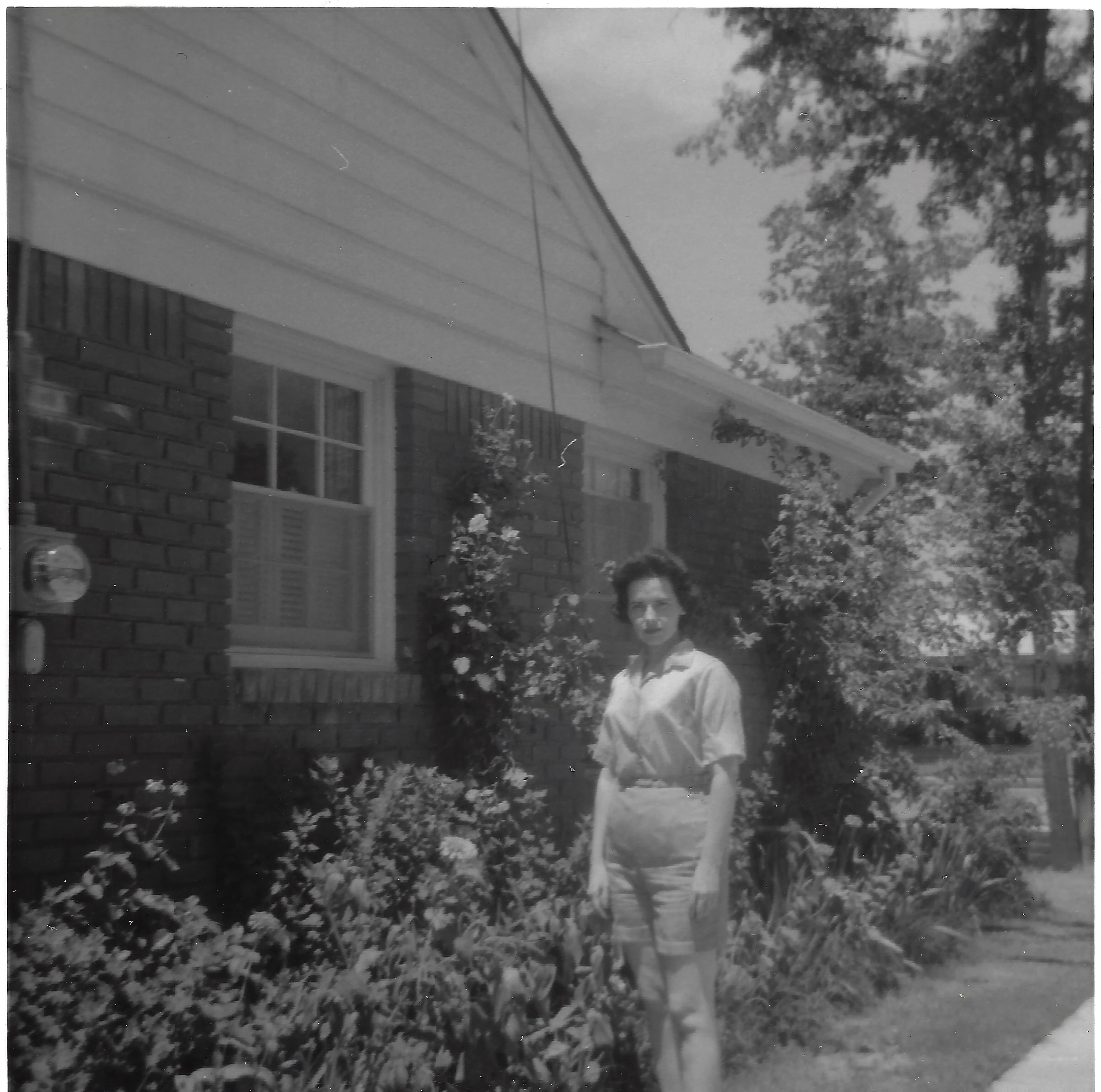 Gruzel (b.1924) in a blouse and shorts standing among flowers beside a brick house, around 1962