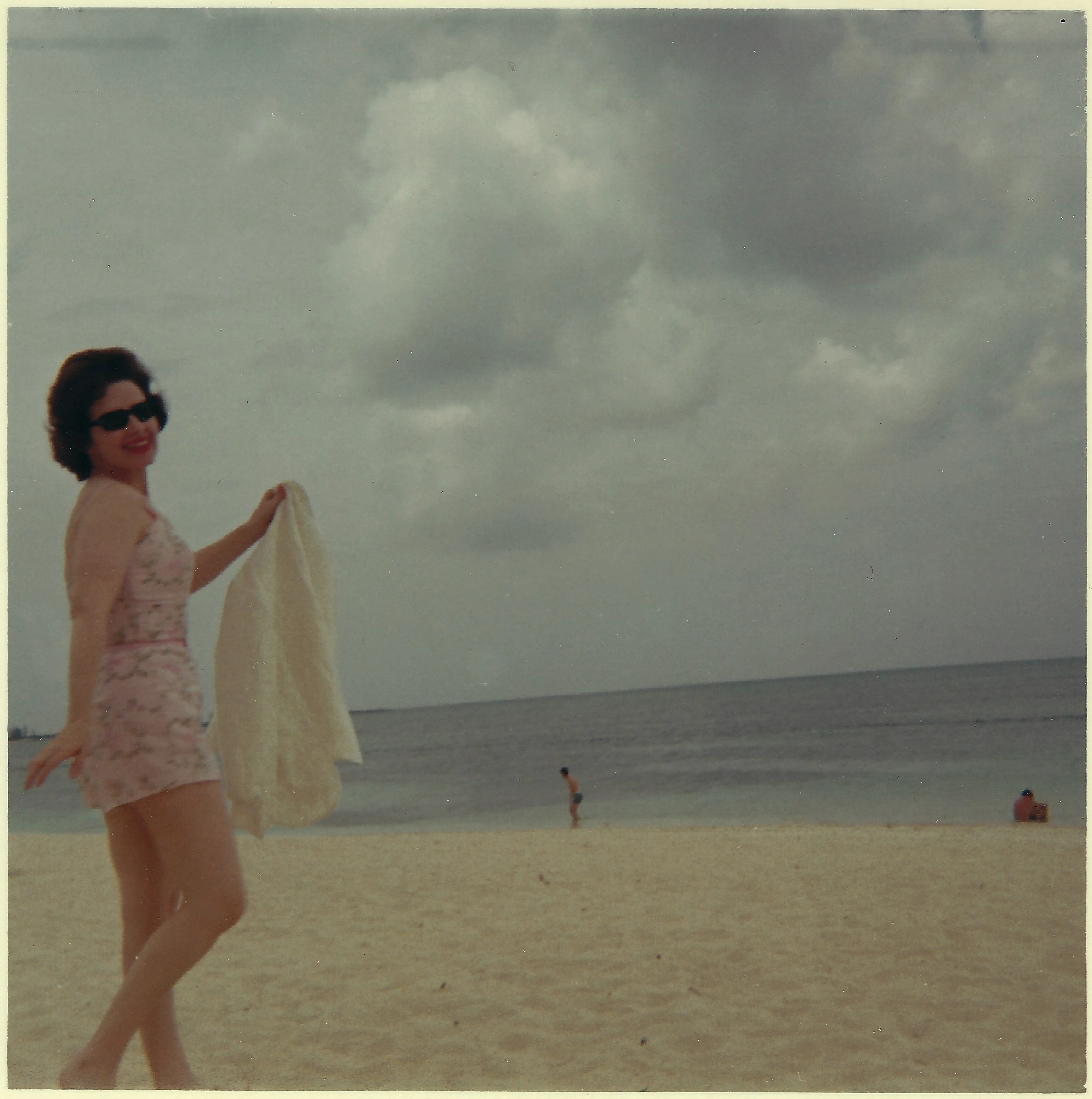 Gruzel (b.1924) in a pink dress and sunglasses holding a scarf on a beach under cloudy skies, around 1965