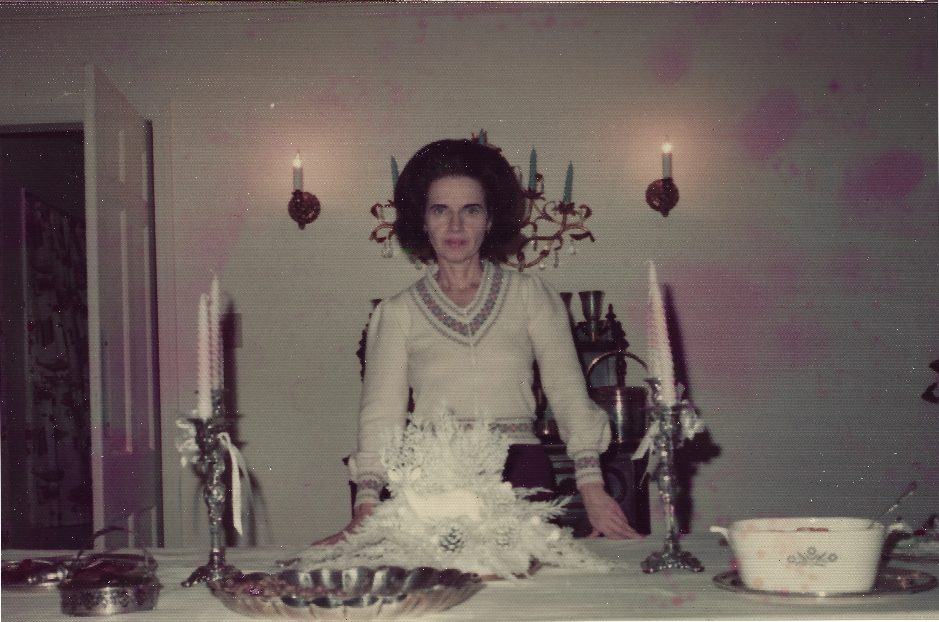 Gruzel (b.1924) in a knit dress standing behind a table set with candelabra and a white floral arrangement, around 1965