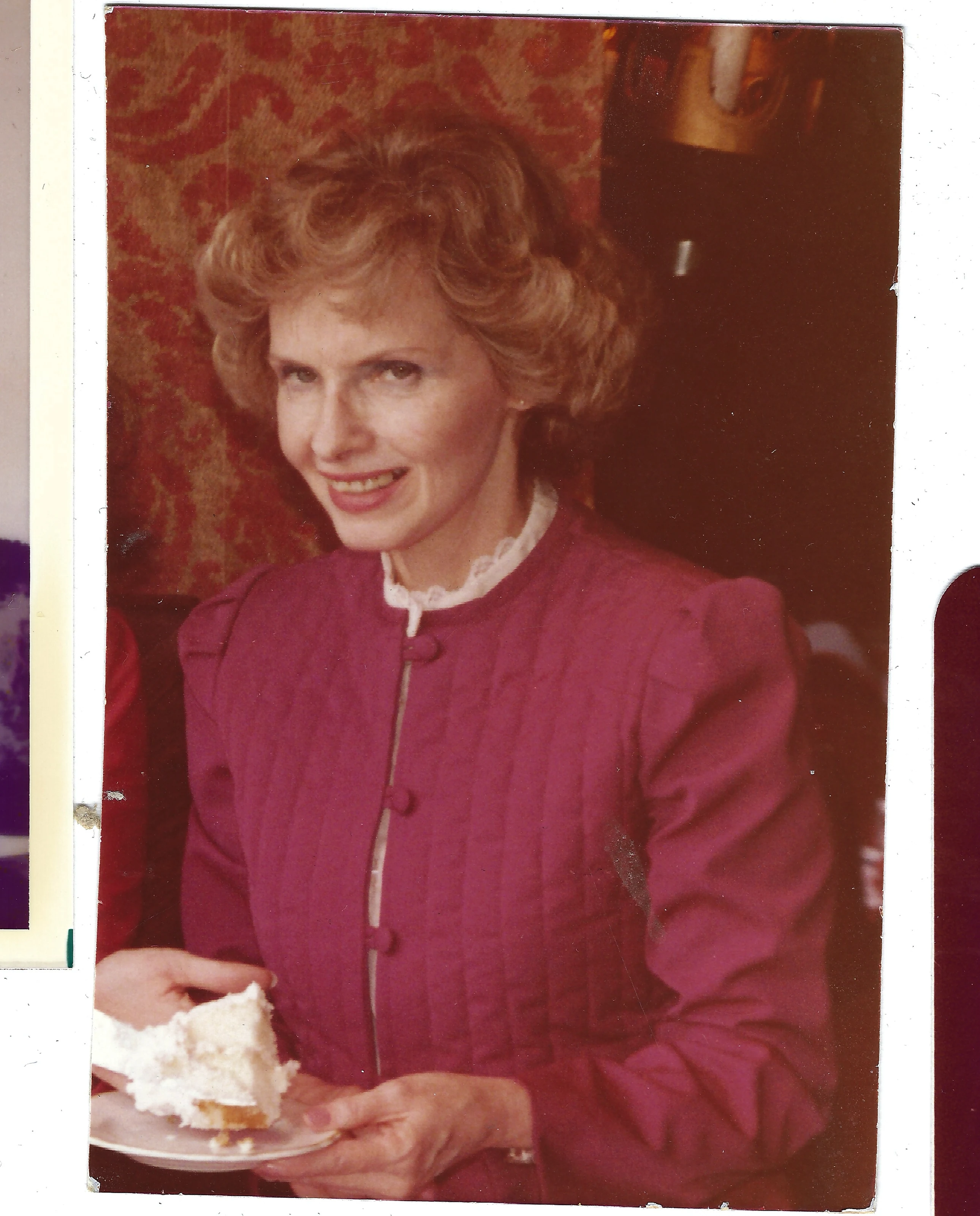 Gruzel (b.1924) in a magenta quilted suit holding a plate of cake at a gathering, around 1983