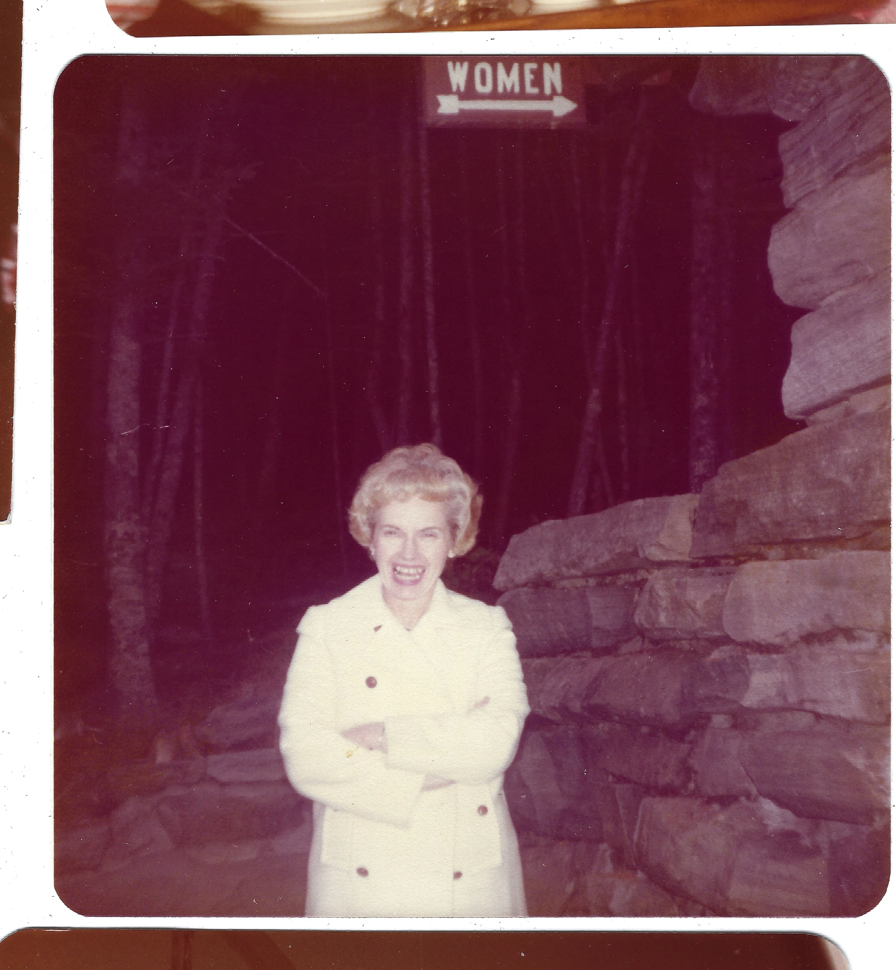 Gruzel (b.1924) in a cream coat with her arms crossed by a stone wall, around 1989