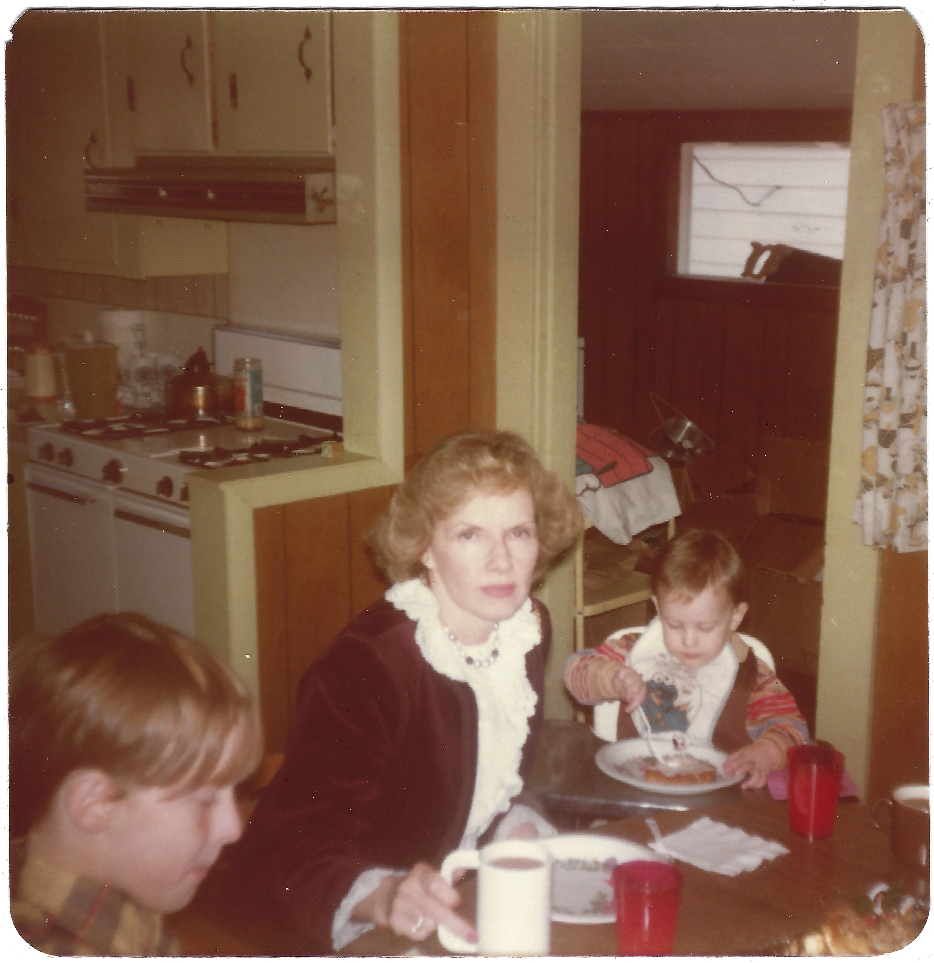 Gruzel at a kitchen table with two grandchildren eating, around 1976
