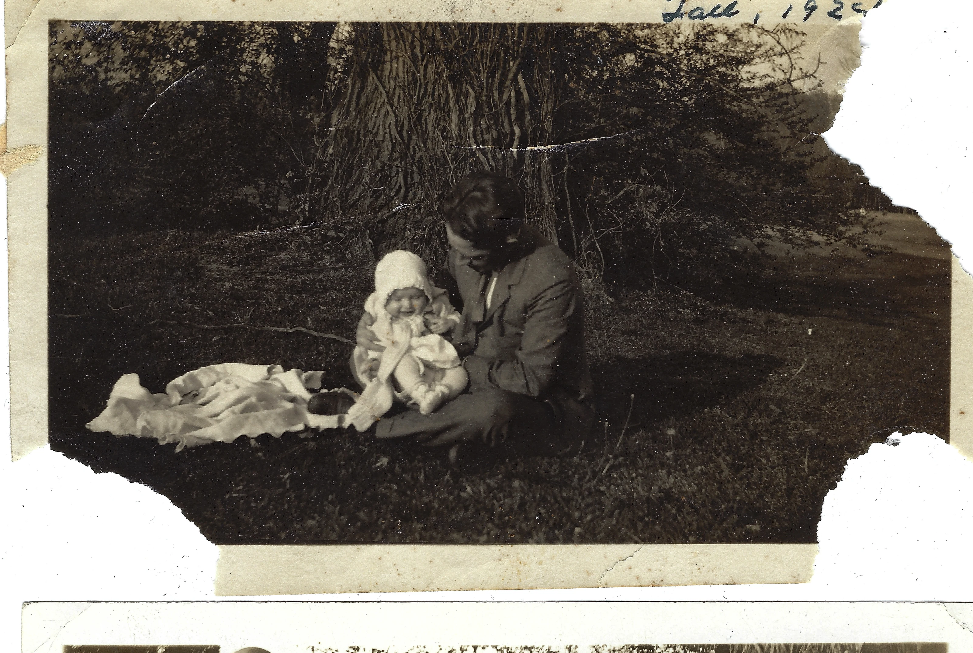 Percy Kerby ('Father Percy') in a suit seated under a tree holding his baby daughter Gruzel in a bonnet on a blanket; the print is noted 'Fall, 1925'