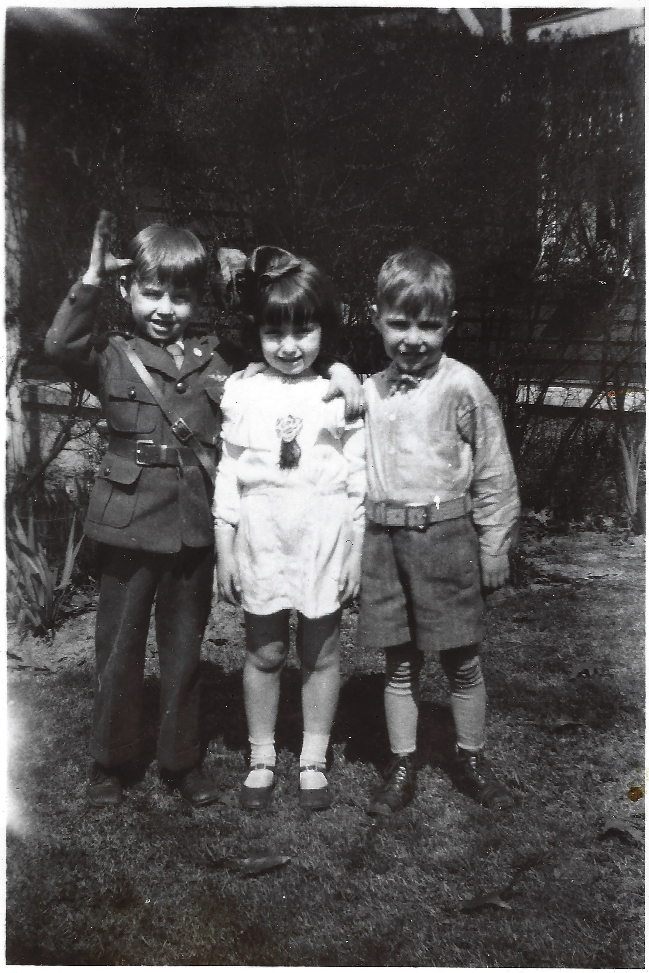Three young children in a yard — a boy in a uniform saluting, a girl in a white dress with a bow, and a boy in shorts — around 1930, from Gruzel's collection