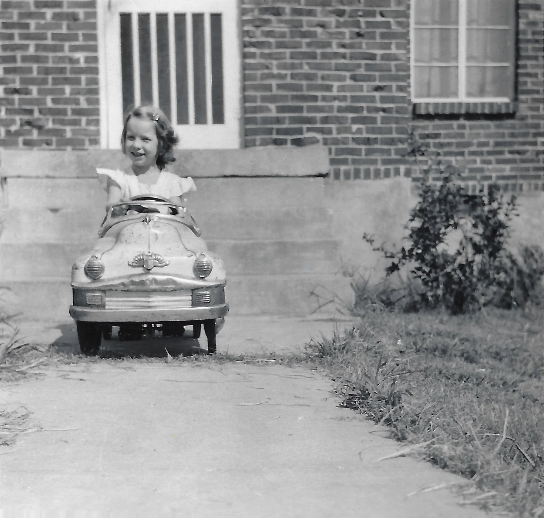 Cacky as a young girl driving a toy pedal car on a front walk before a brick house, around 1953