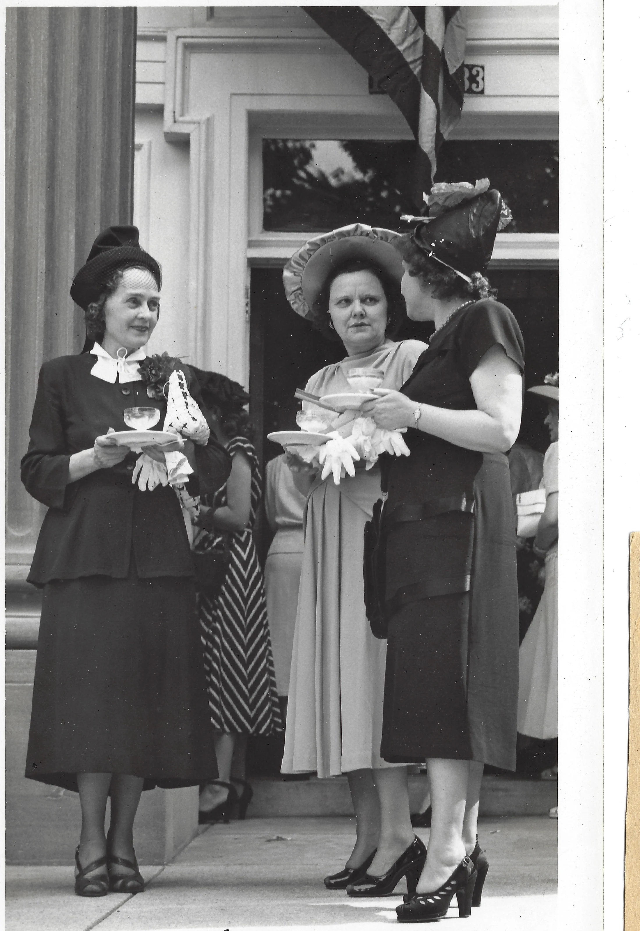 Mama Percy (Catherine Fleming) among women in 1940s hats, gloves and platform heels holding teacups at an outdoor reception, around 1944
