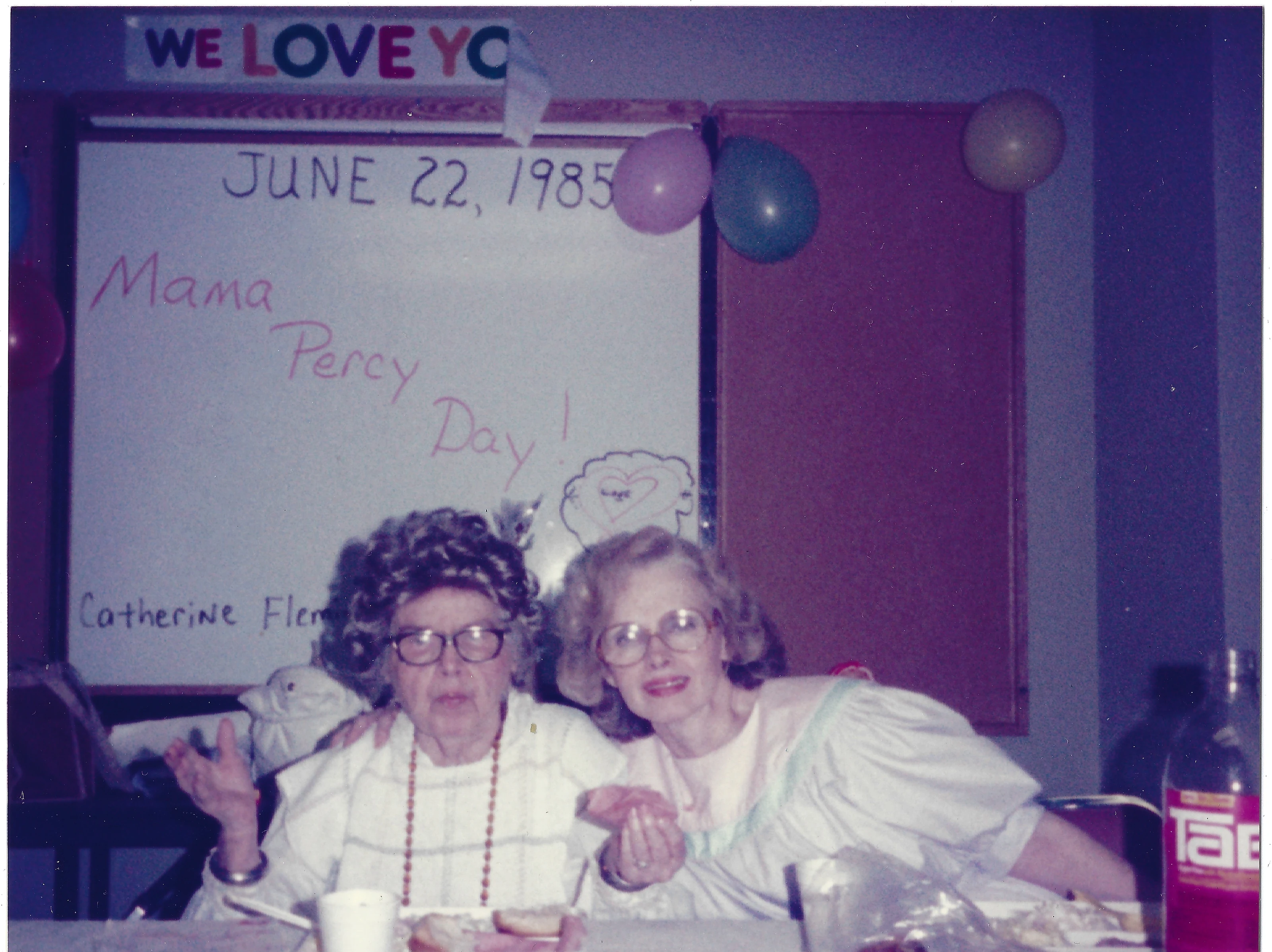 Mama Percy (Catherine Fleming) and Gruzel at the "Mama Percy Day" celebration in front of a hand-lettered banner dated June 22, 1985