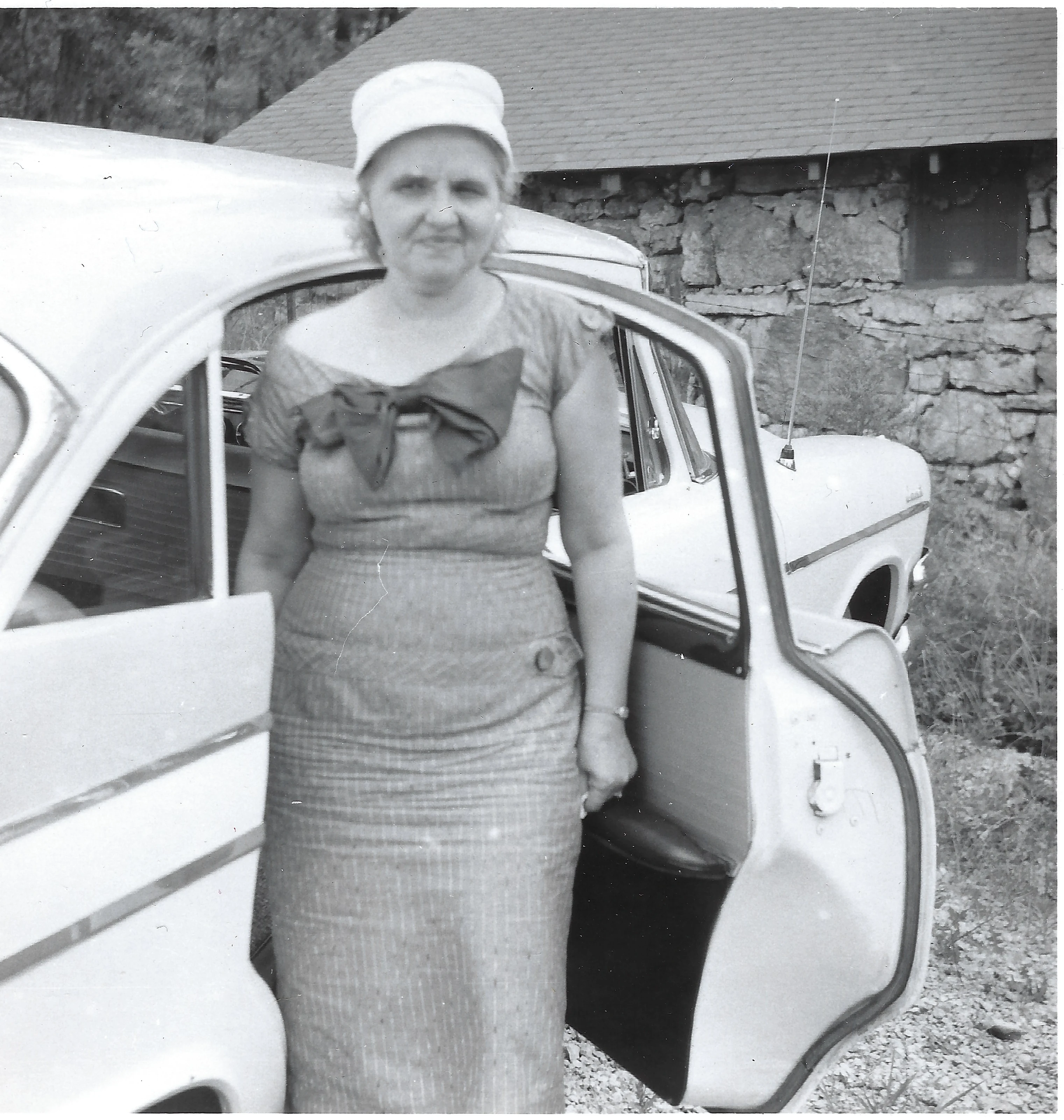 Mama Percy (Catherine Fleming) in a cap and bow-trimmed dress standing beside a brand-new car; the print is lab-stamped May 1958