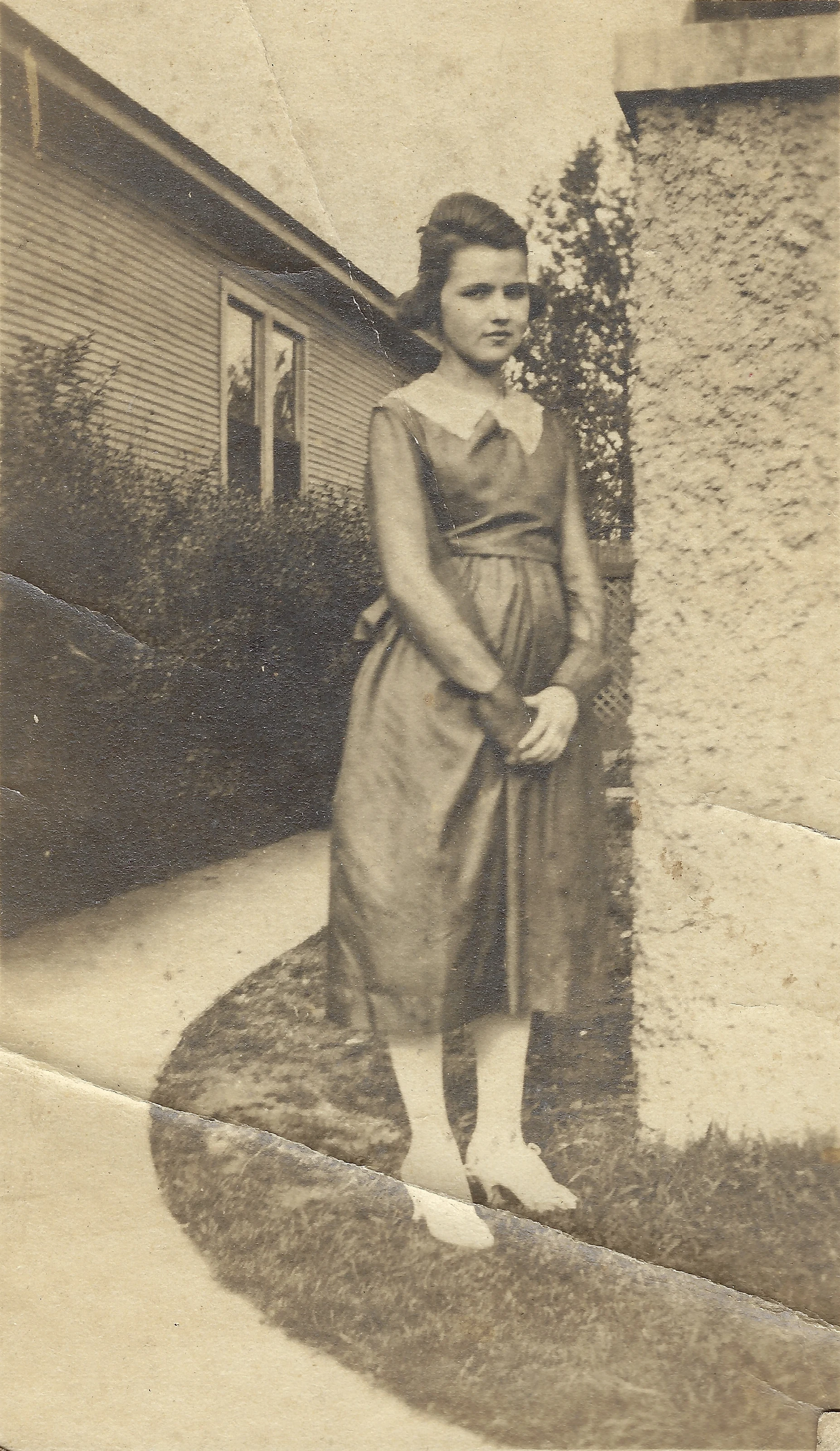 Mama Percy (Catherine Fleming) as a young woman in a 1920s drop-waist dress on a garden walk beside a clapboard house, around 1921