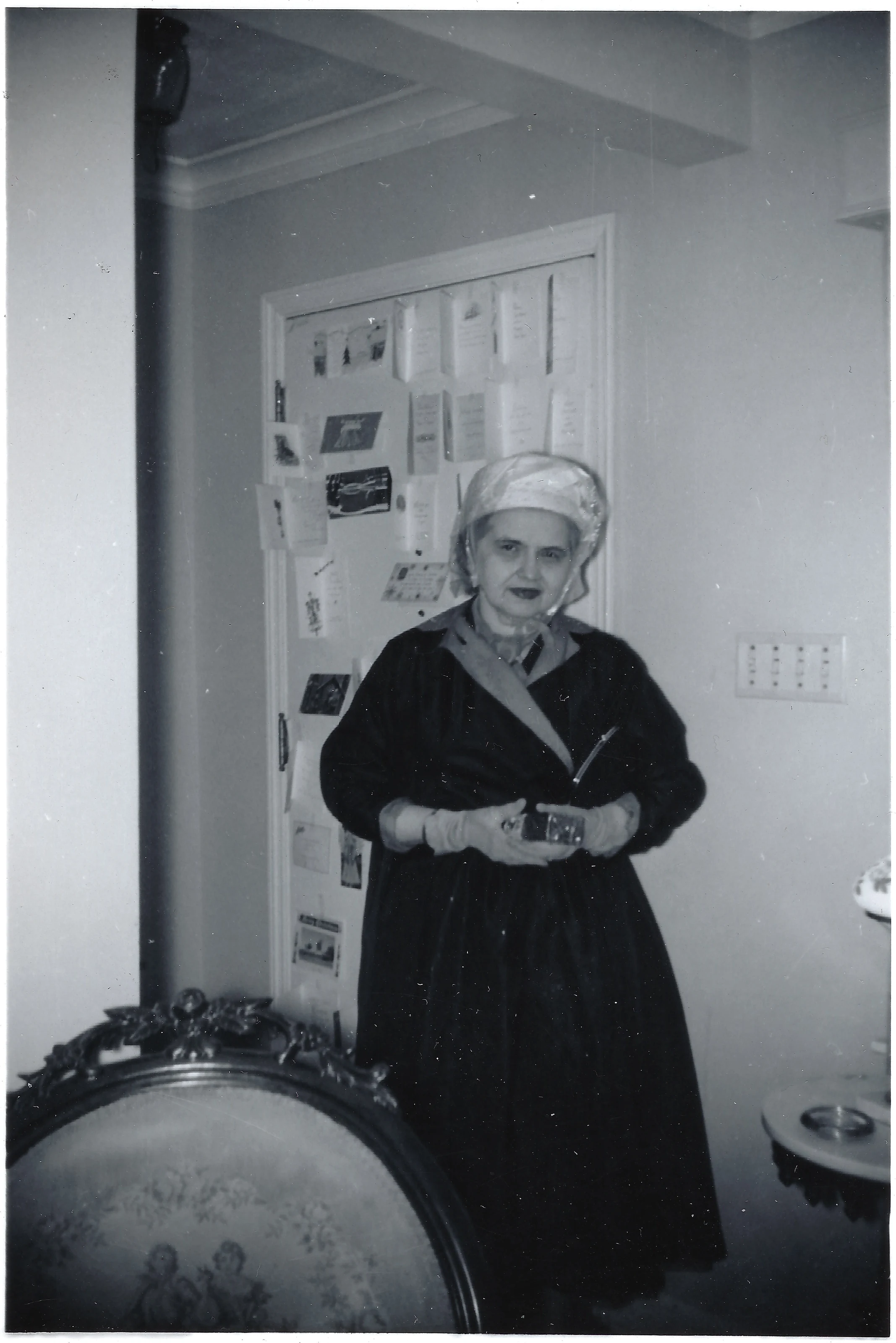 Mama Percy (Catherine Fleming) in a hat and coat by a display of greeting cards, standing in a room, around 1960