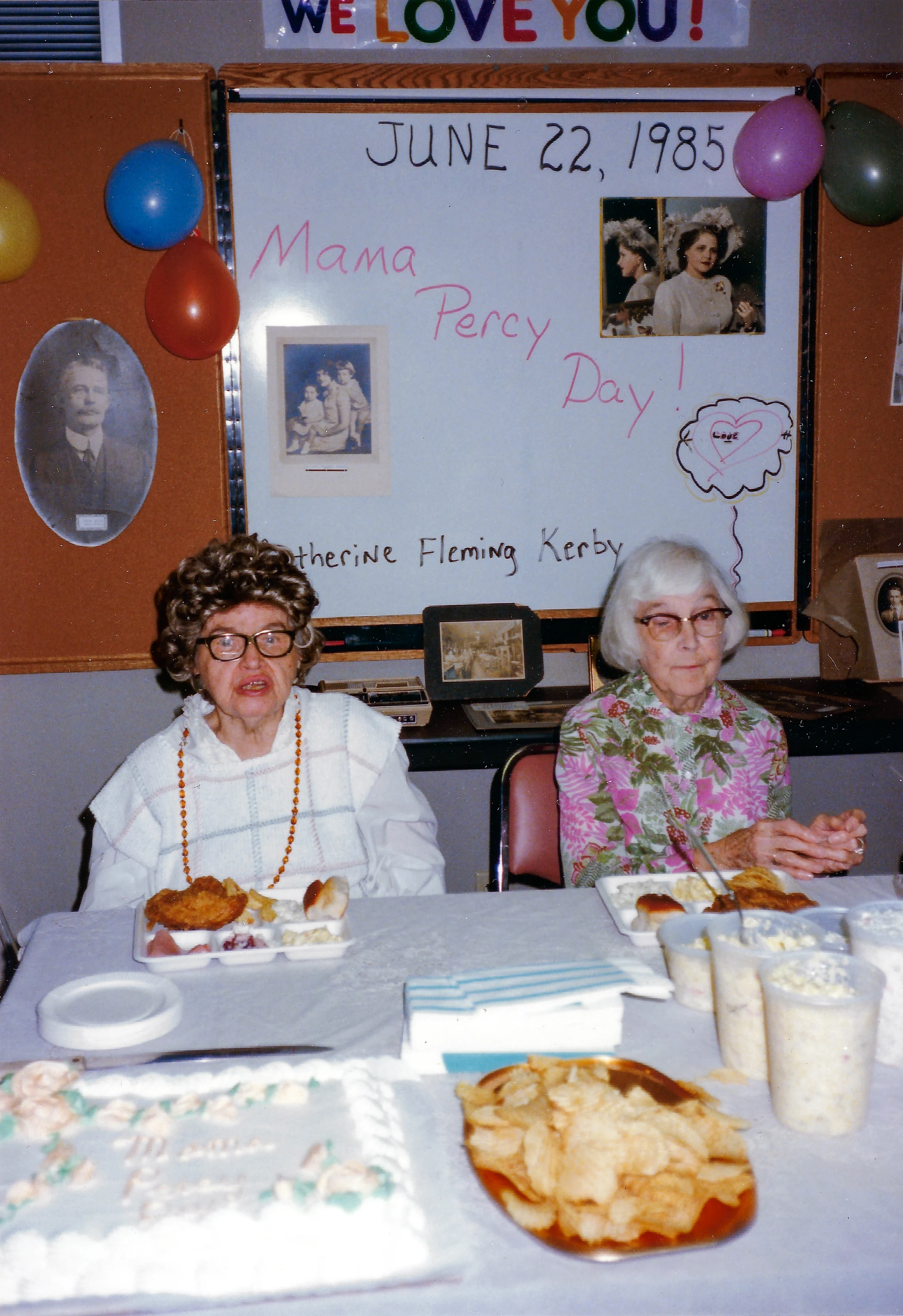 Two women eating lunch beneath the Mama Percy Day sign on June 22, 1985