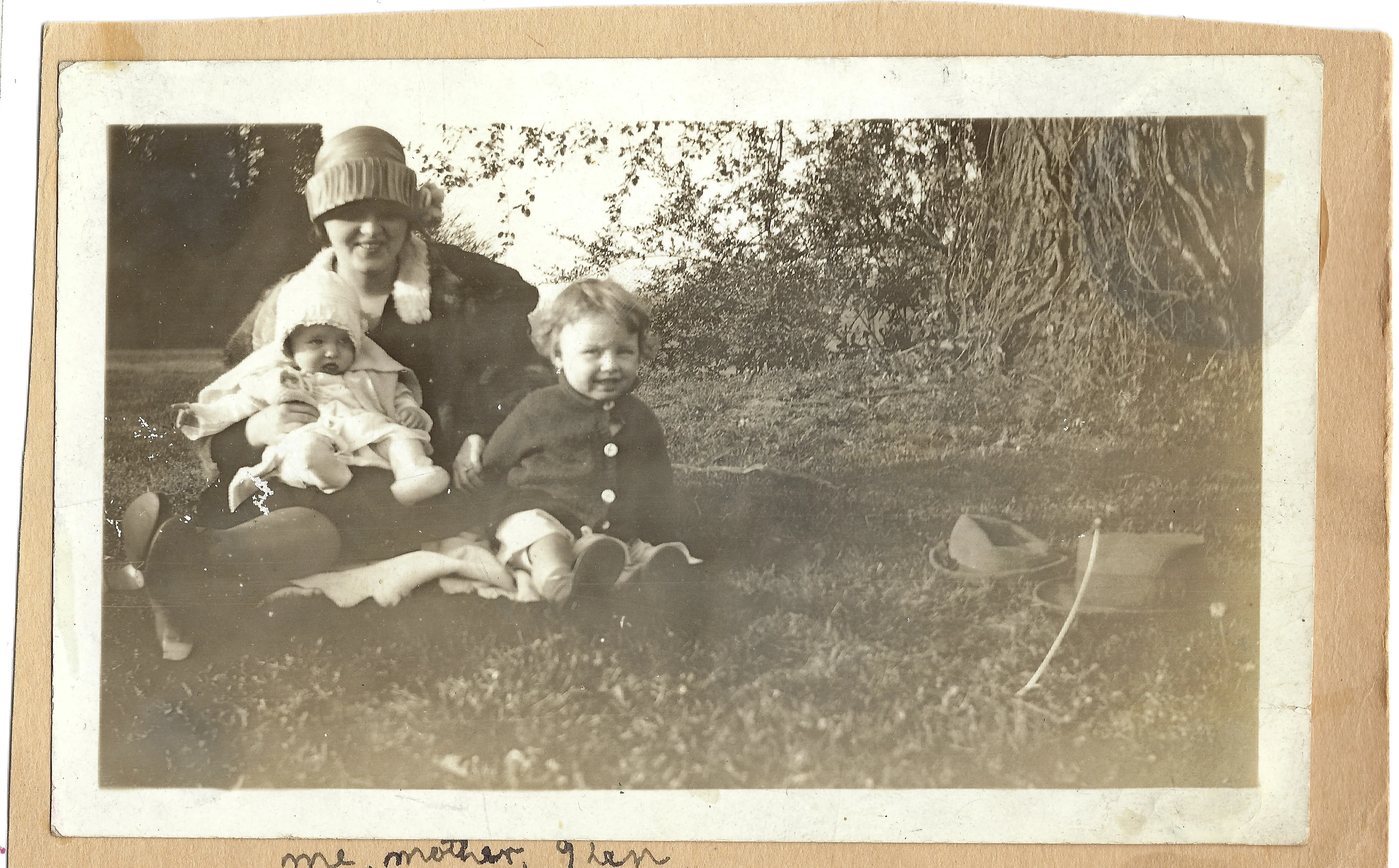 A woman in a cloche hat seated on the grass holding a baby beside a young boy; captioned in the album 'Me, Mother & Ken' — baby Gruzel with her mother Mama Percy and her brother Kenny, around 1925