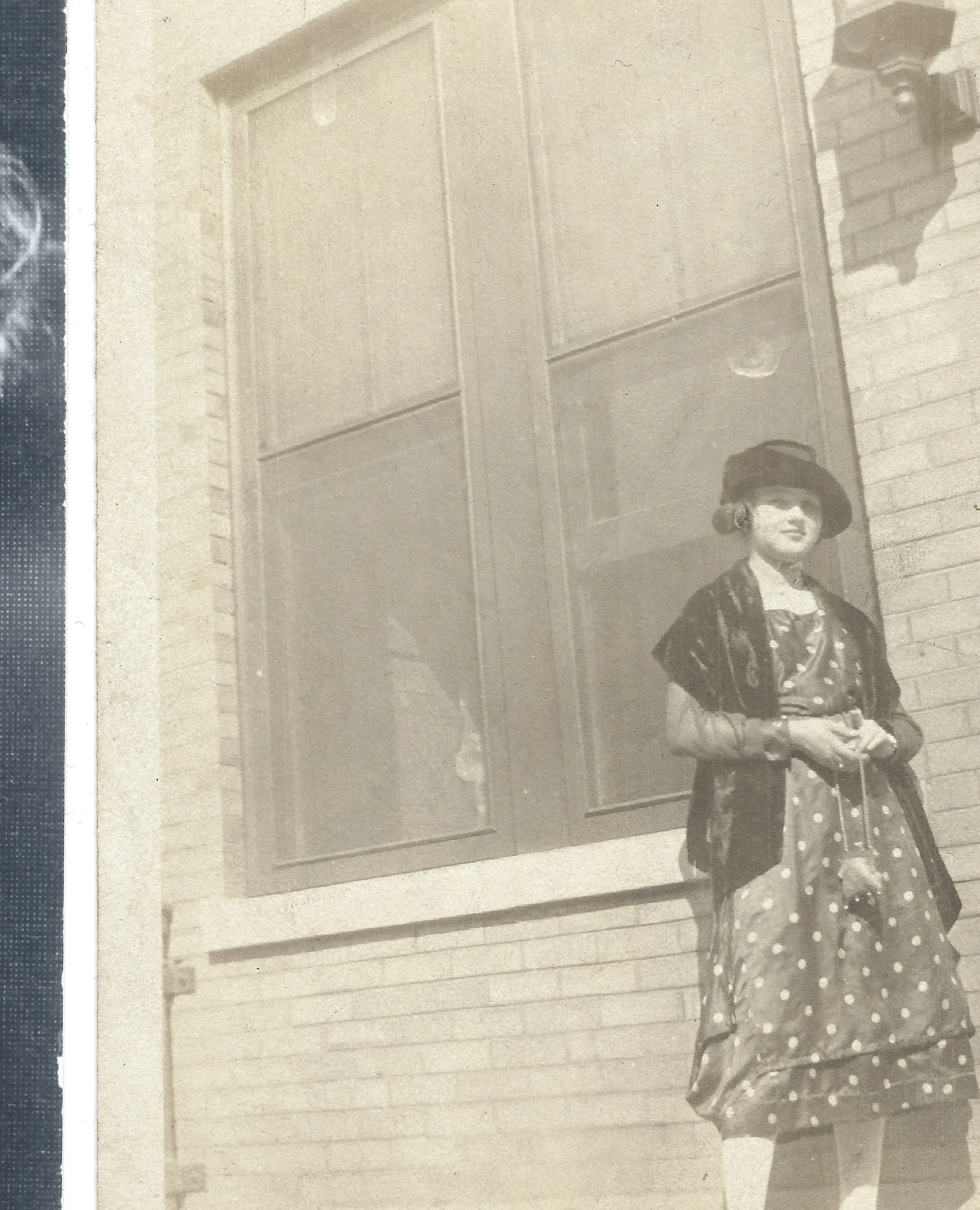 Mama Percy (Catherine Fleming) in a polka-dot dress and hat with a velvet capelet, standing by a brick building, around 1928